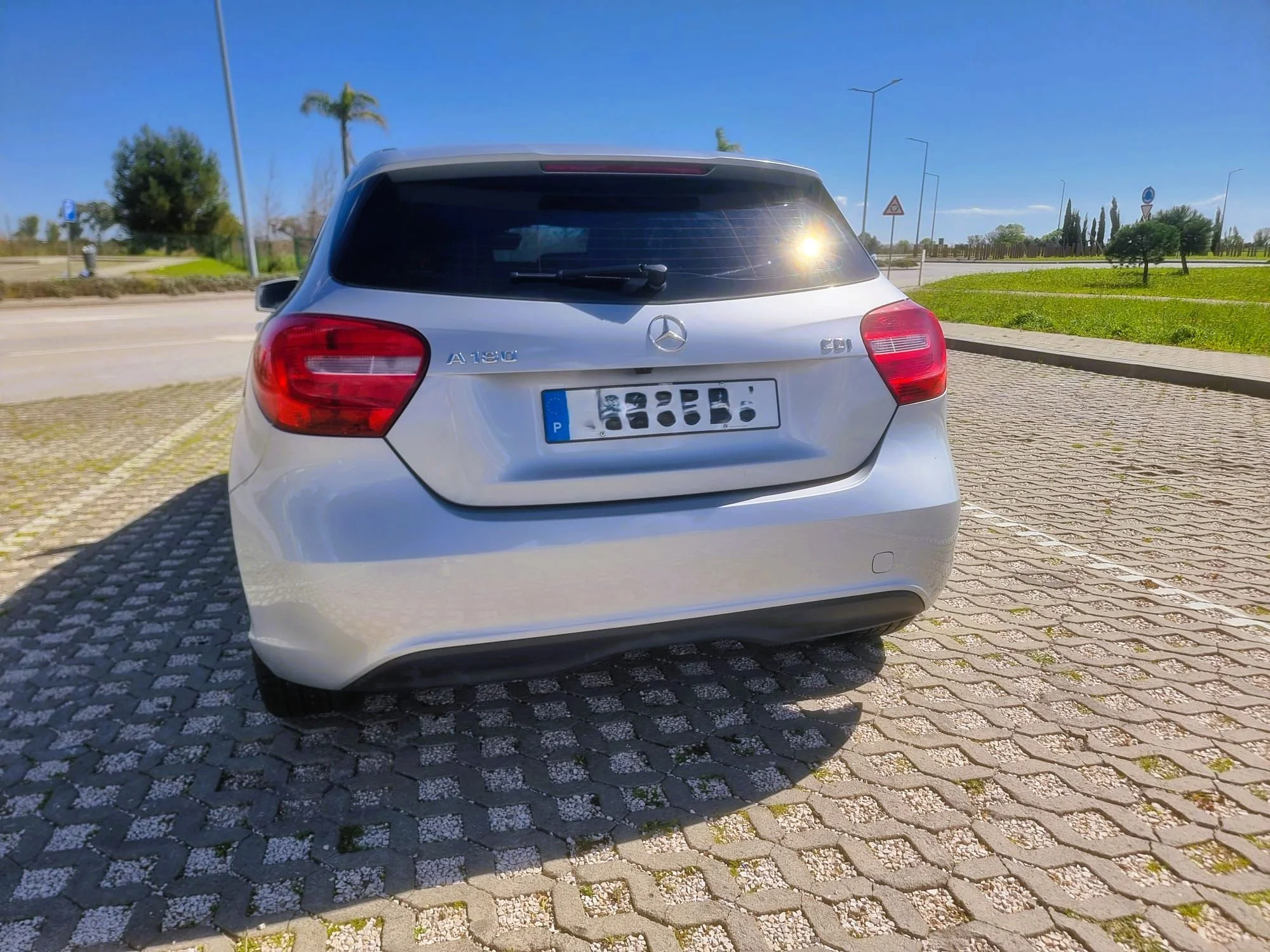 Carro prata estacionado em uma área de estacionamento com piso de blocos de cimento, sob céu claro e com árvores ao fundo.