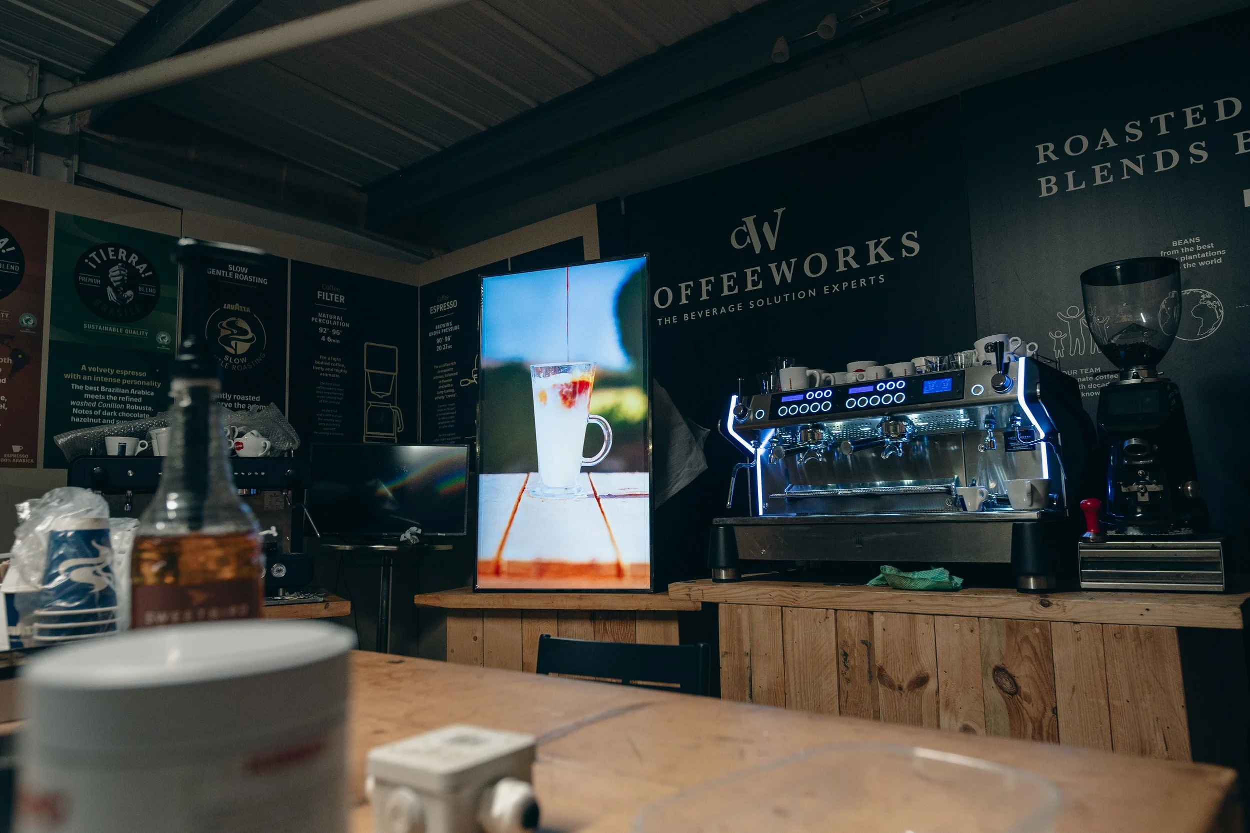 The image shows a coffee shop counter with a visible espresso machine, cups, and signage about coffee beans and roasting. There is a large digital advertisement screen behind the counter displaying a glass of iced coffee. The environment has a rustic, wooden decor.