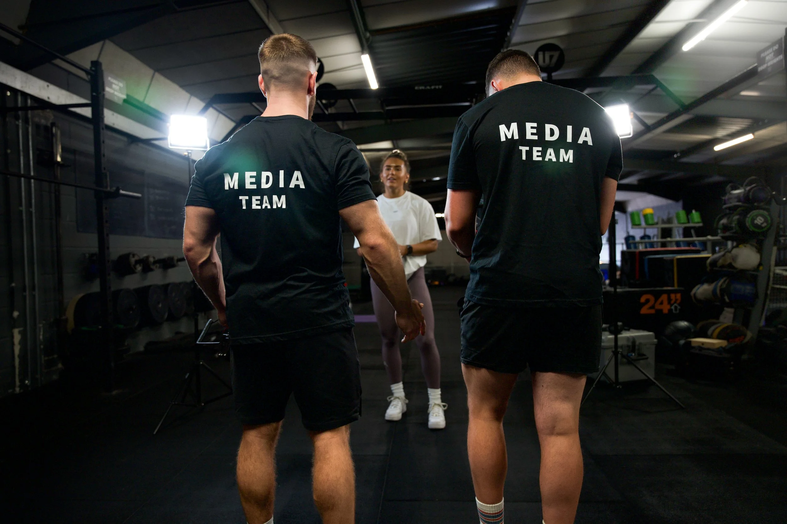 Two men wearing black t-shirts with 'MEDIA TEAM' written on the back stand in a gym, talking while facing a woman who appears to be giving instructions or having a conversation. The gym has workout equipment, weights, and bright lighting.