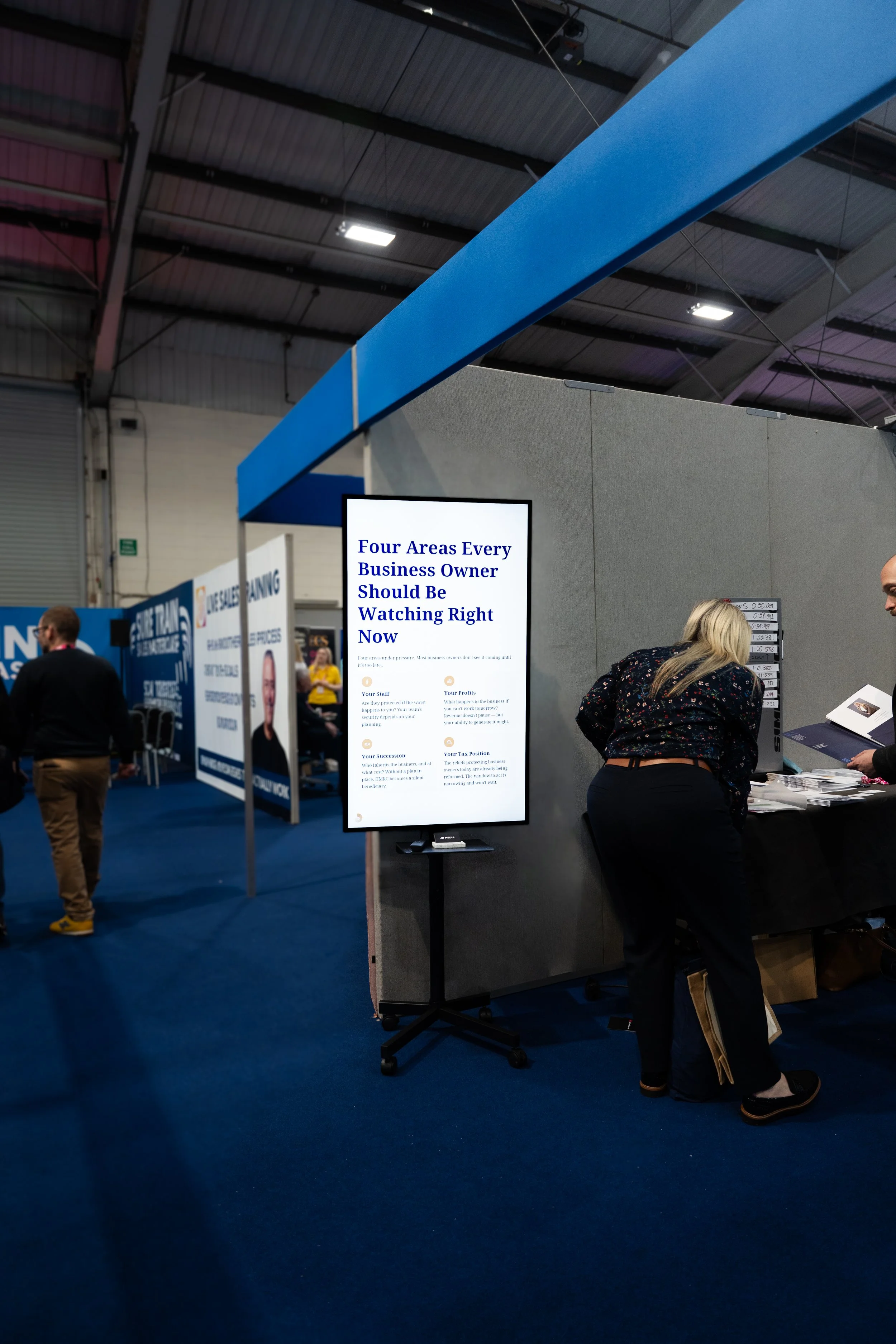 People at a trade show booth, with a large vertical digital screen displaying text about four areas every business owner should watch.