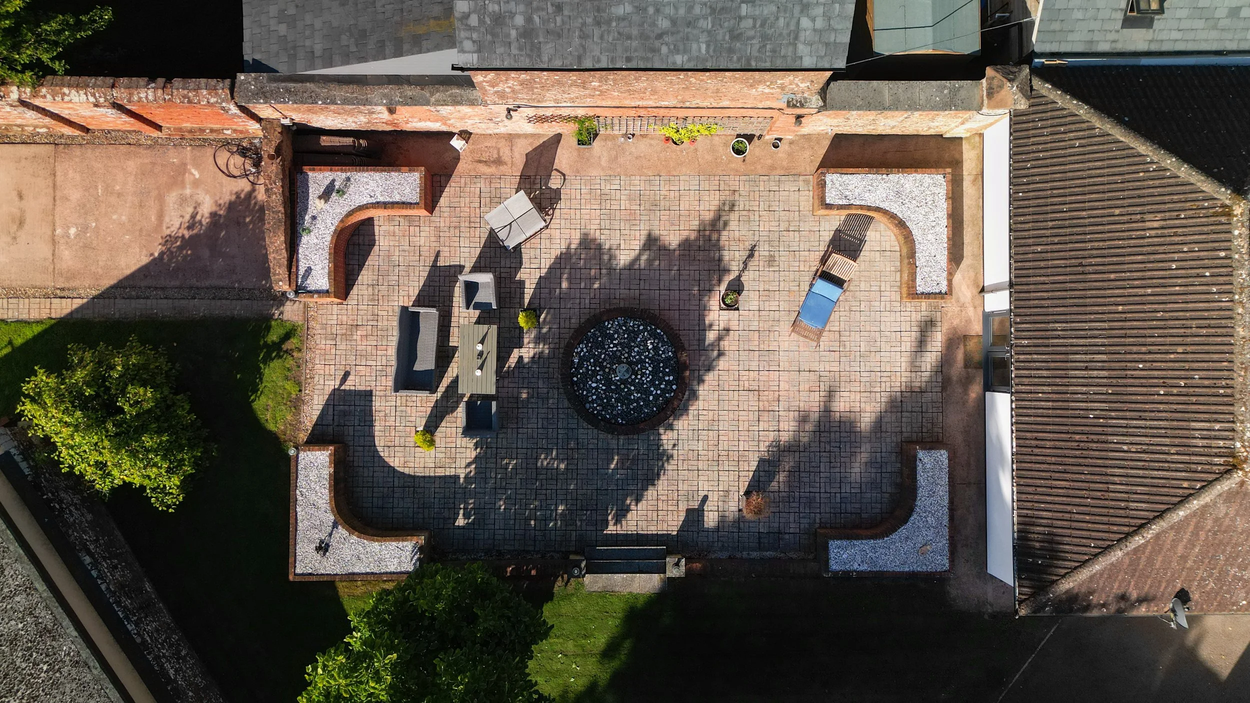 Overhead view of a backyard patio with brick flooring, outdoor furniture including a table with chairs, a bench, a circular decorative rock feature, potted plants, and surrounding brick and stone walls with some plants and a tree.