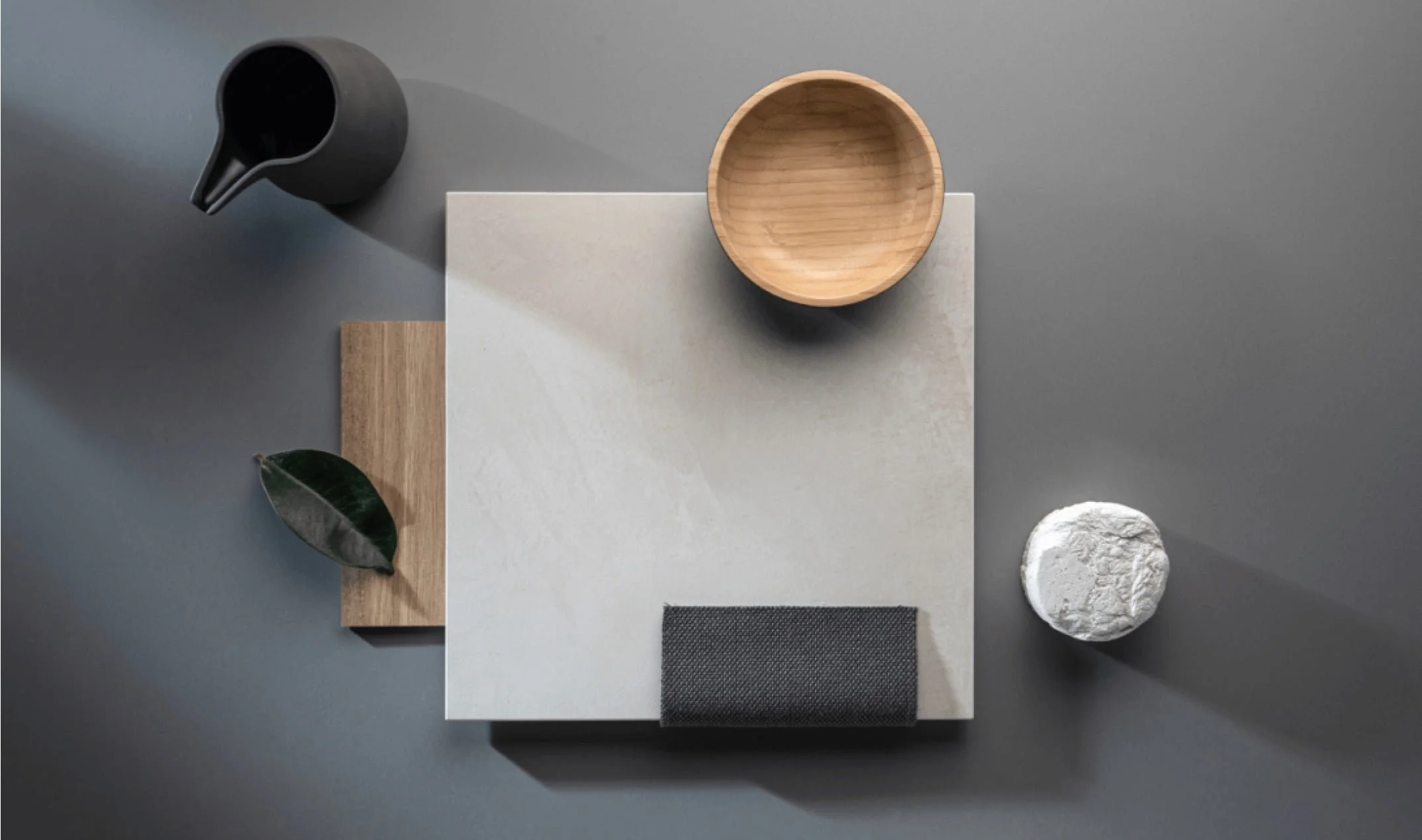 Flat lay of various objects on a gray background, including a black pitcher, a wooden bowl, a black vase, a rectangular black piece, a light wood board, a green leaf, and a white textured stone.