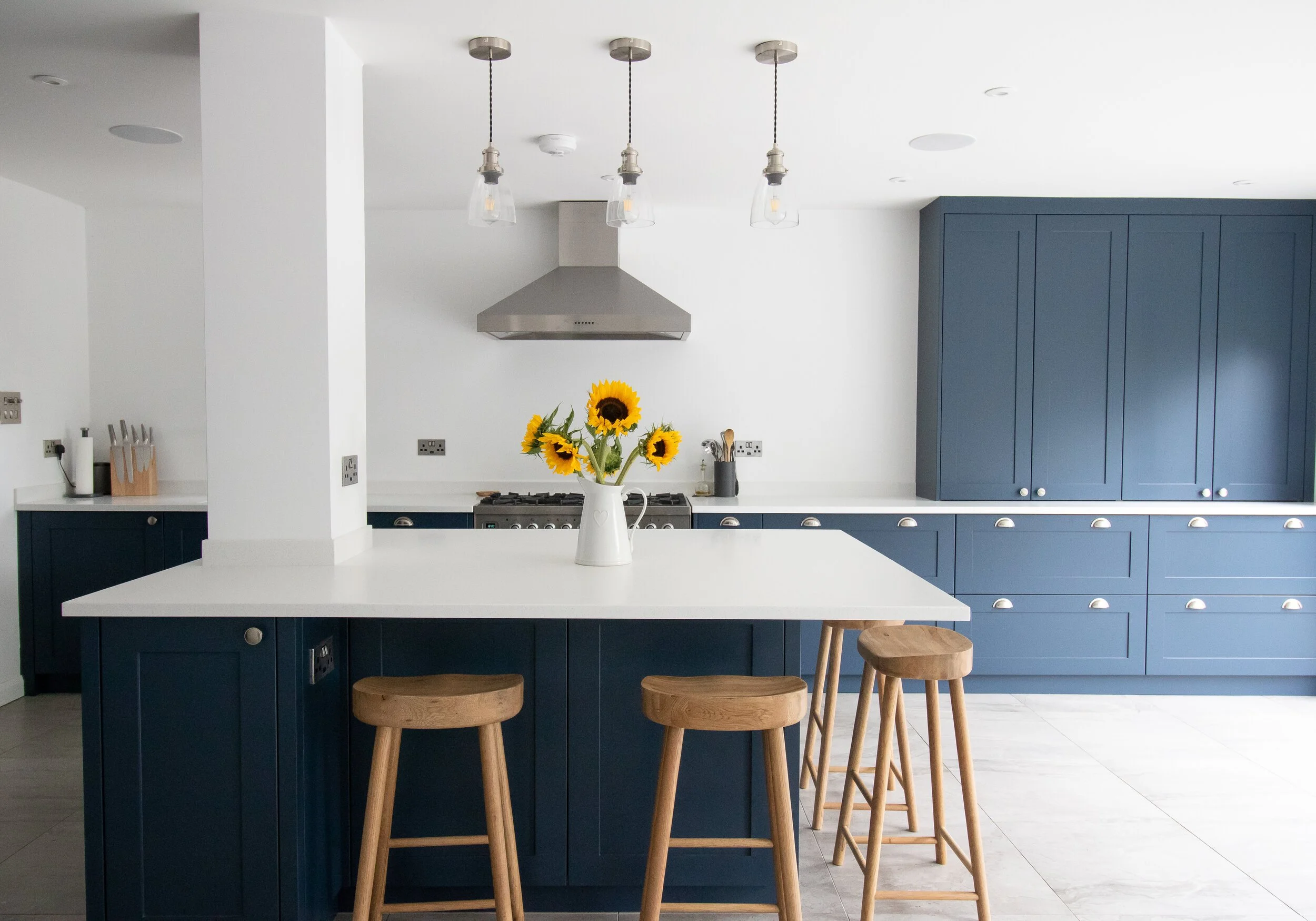 Modern kitchen with blue cabinets, white countertops, a white island with wooden stools, a sunflower bouquet in a white pitcher, stainless steel hood, and pendant lights.