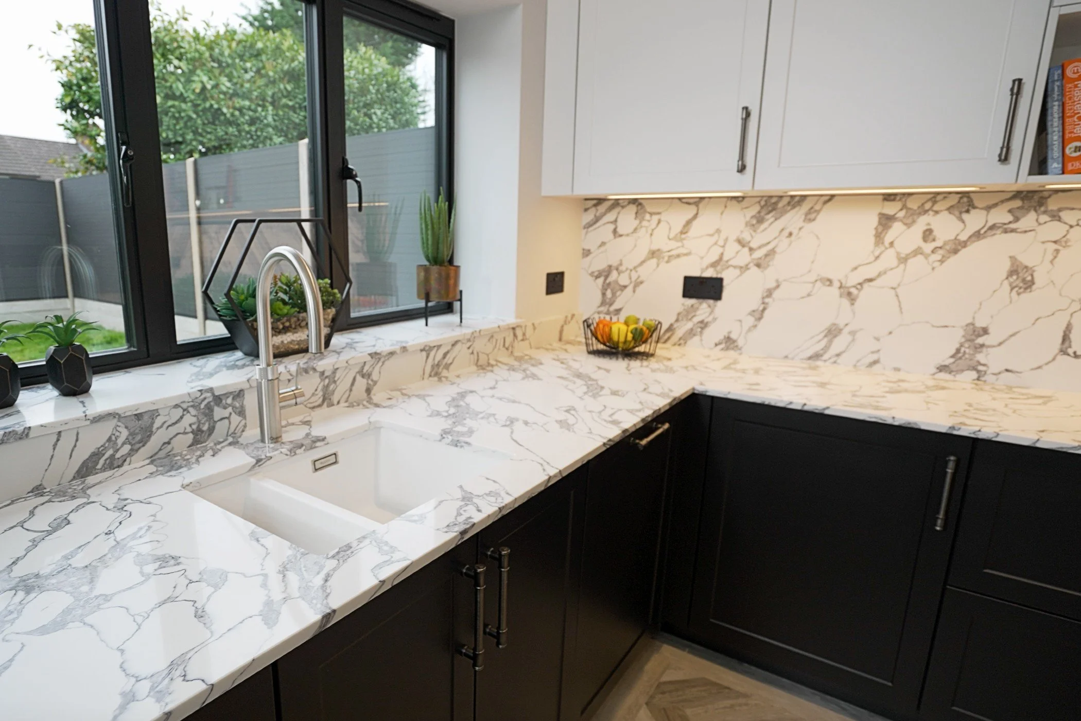 Modern kitchen with white and black cabinetry, marble countertops, and a sink under a large window with a view of a backyard.