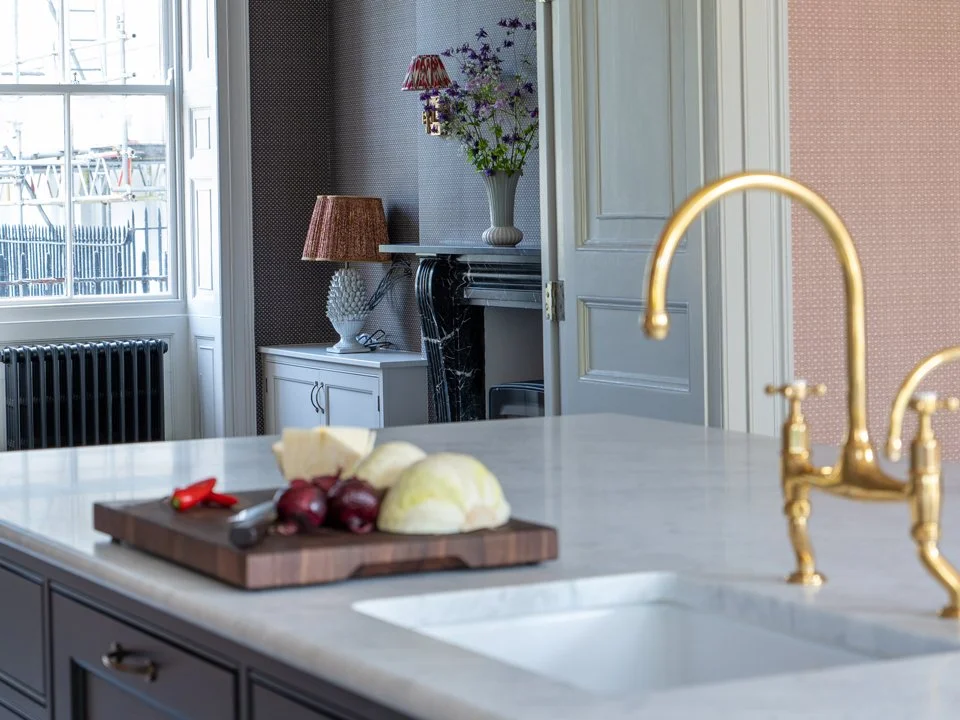 A kitchen countertop with a cutting board, vegetables, and a gold faucet with a white sink. In the background, there is a corner with a fireplace, a small white cabinet, a table lamp, and a large vase with purple flowers. A window shows an outdoor view.