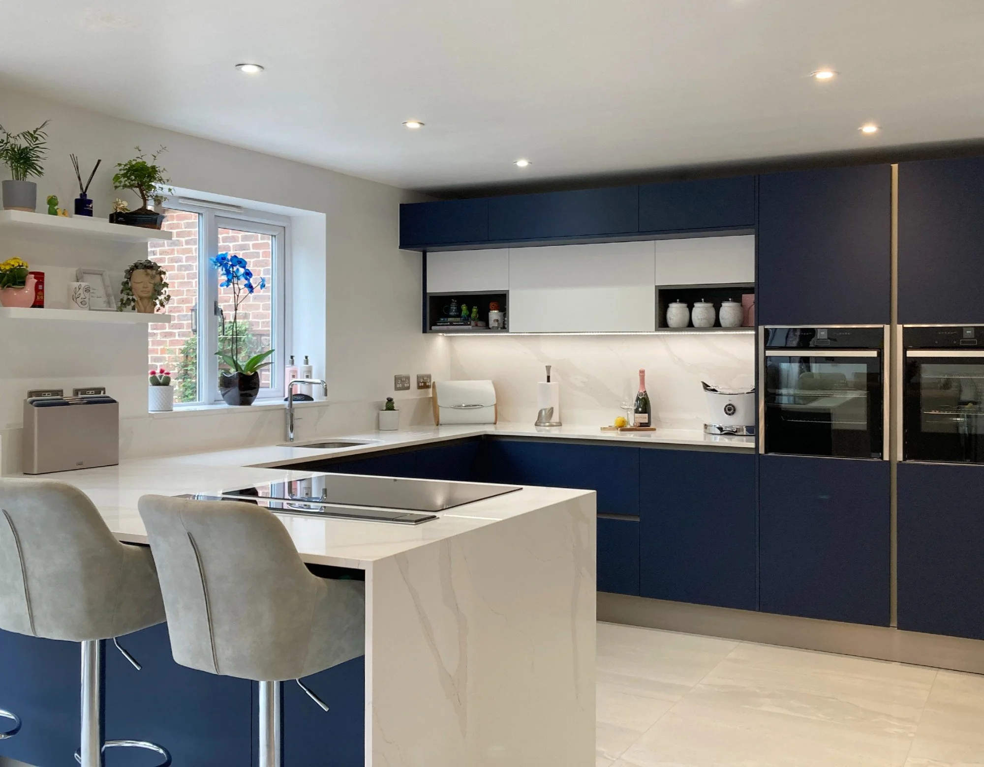Modern kitchen with white and navy blue cabinets, marble countertops, window with brick wall outside, beige chairs at white island, small appliances, and plants.