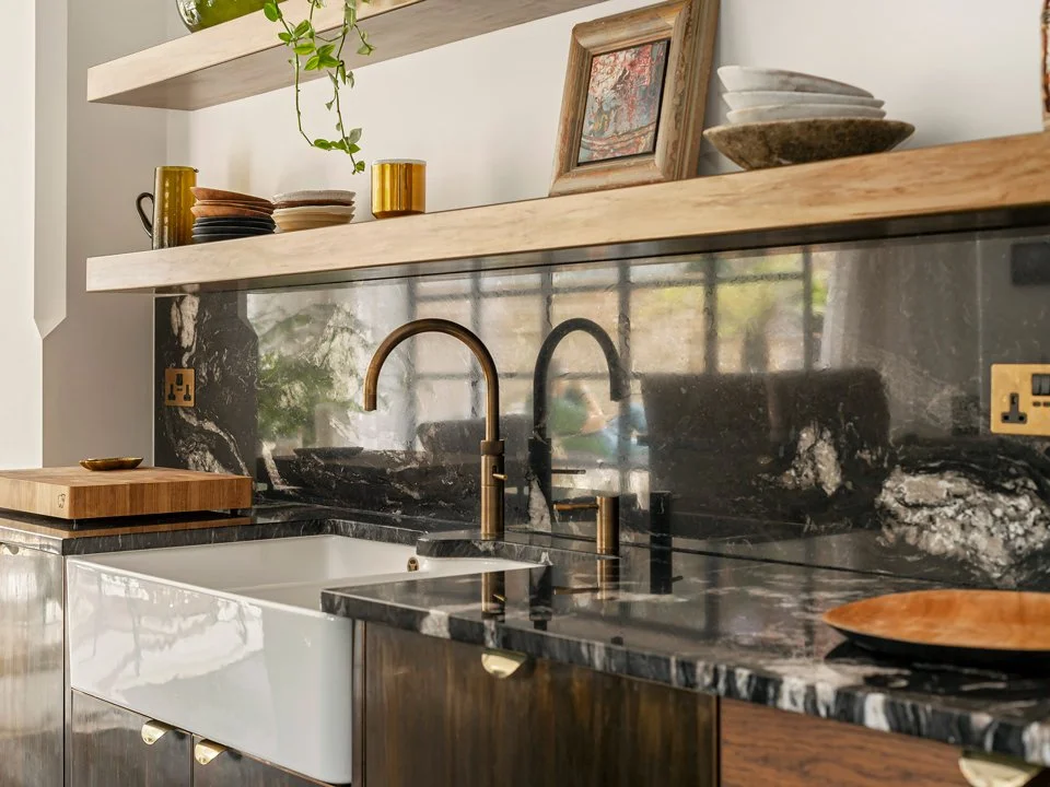 Modern kitchen with black marble countertops, brass fixtures, white farmhouse sink, and wooden open shelves displaying dishes and decorative items.