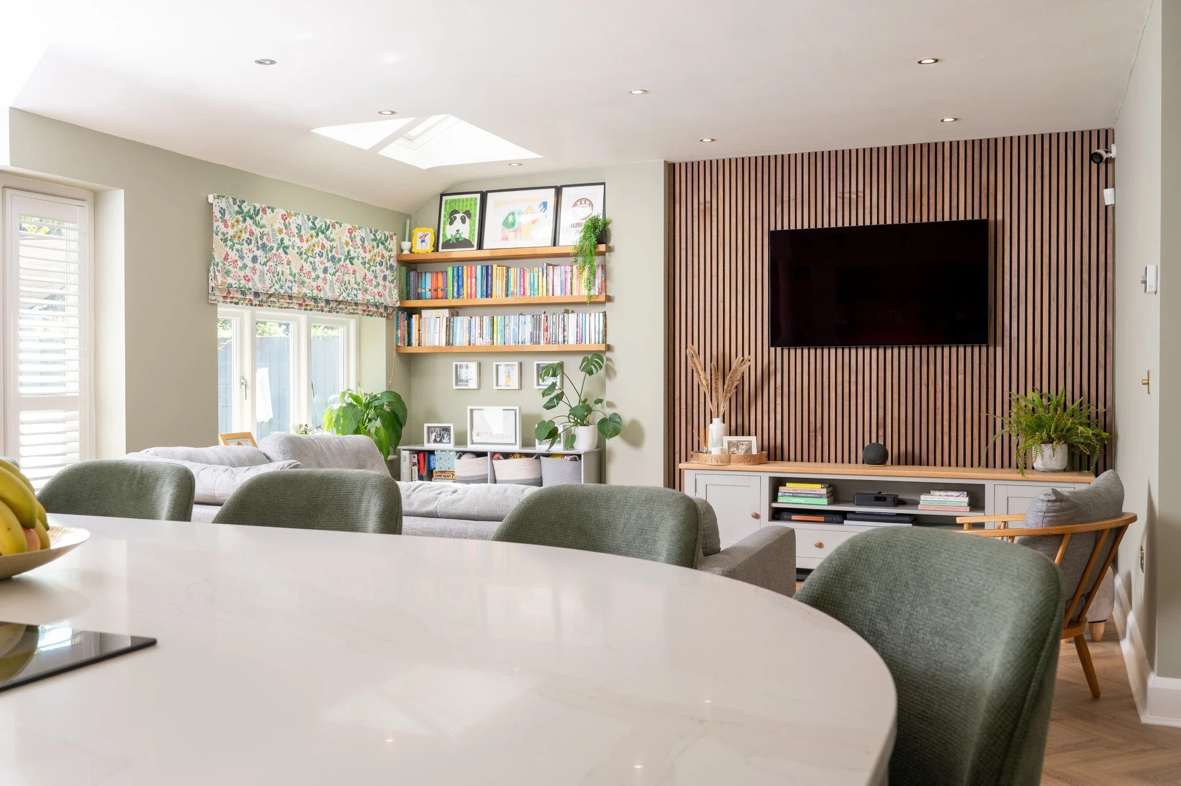 Bright living room with gray couches, a large flat-screen TV on a wooden slat wall, bookshelves filled with books and framed photos, and green indoor plants, with a large window and skylight letting in natural light.