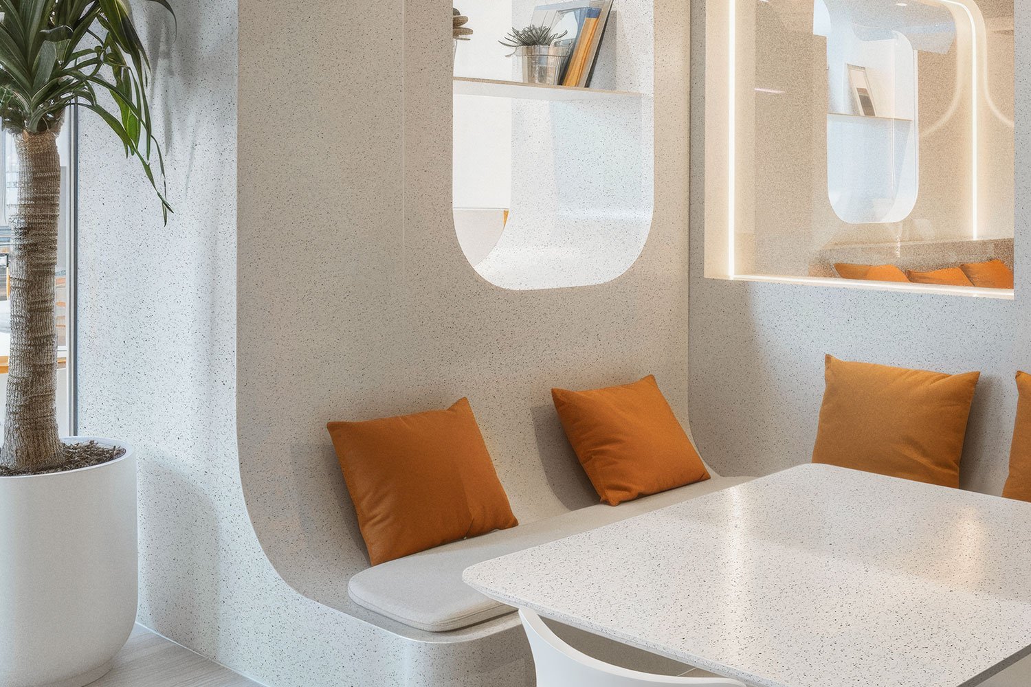 Modern interior with a white speckled table, orange cushions on a built-in curved bench, and a potted plant near a window.