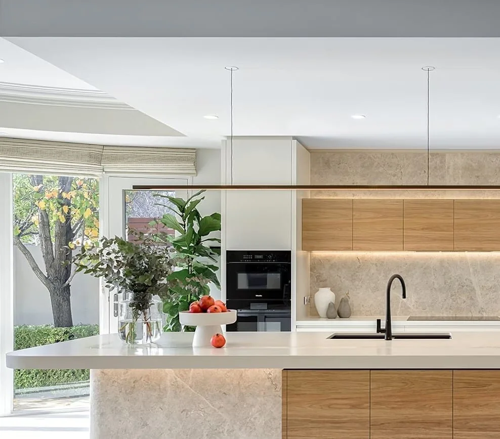 Modern kitchen with white countertops, wooden cabinets, black faucet, and built-in oven. There are potted plants and a vase with flowers on the countertop, and a large window showing trees outside.
