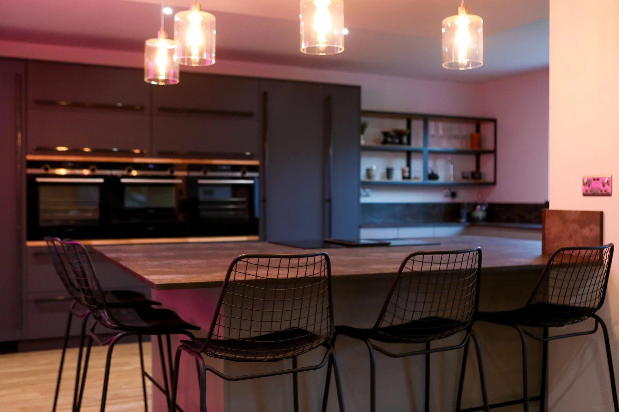 Modern kitchen with a wooden island counter, black wire chairs, gray cabinets, open shelving, and hanging pendant lights.