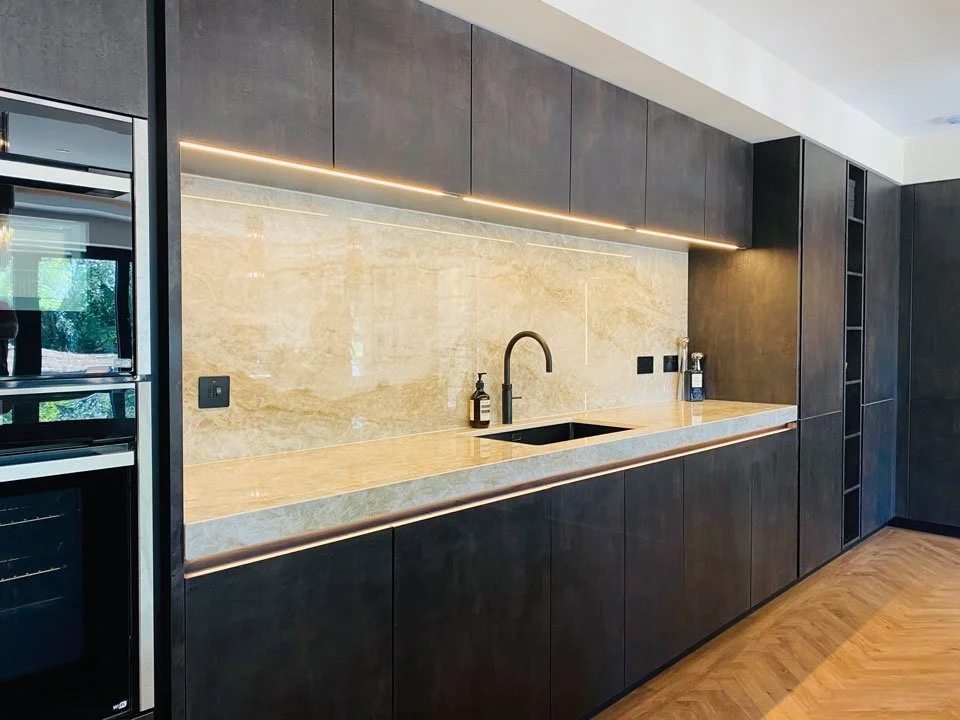 Modern kitchen with dark cabinetry, a beige marble backsplash, and a wooden countertop. Features a black sink and a black gooseneck faucet, with built-in lighting underneath the upper cabinets.