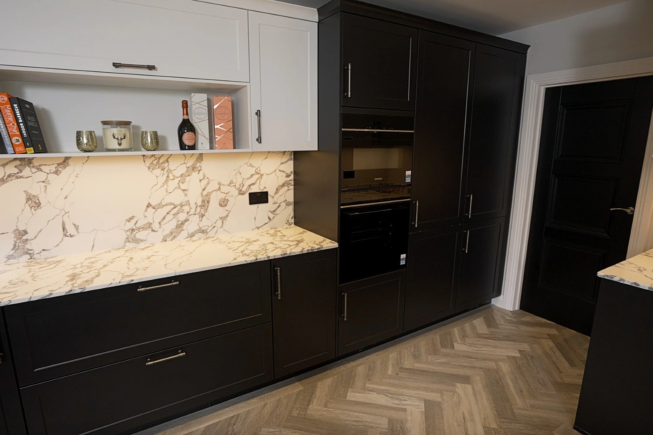 Modern kitchen with black cabinets, white and gold marble countertops, and a marble backsplash. Open shelf with books, candles, and decorative items. Wooden herringbone flooring.
