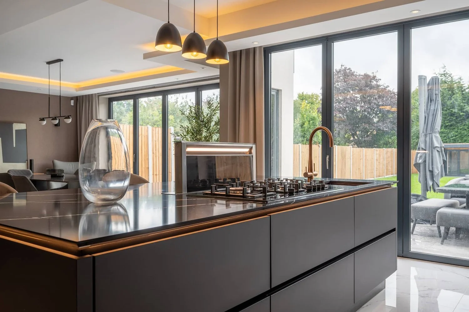 Modern kitchen with a black island, built-in stove, gold faucet, large windows, sliding glass door, outdoor patio with seating and an umbrella, and contemporary lighting fixtures.