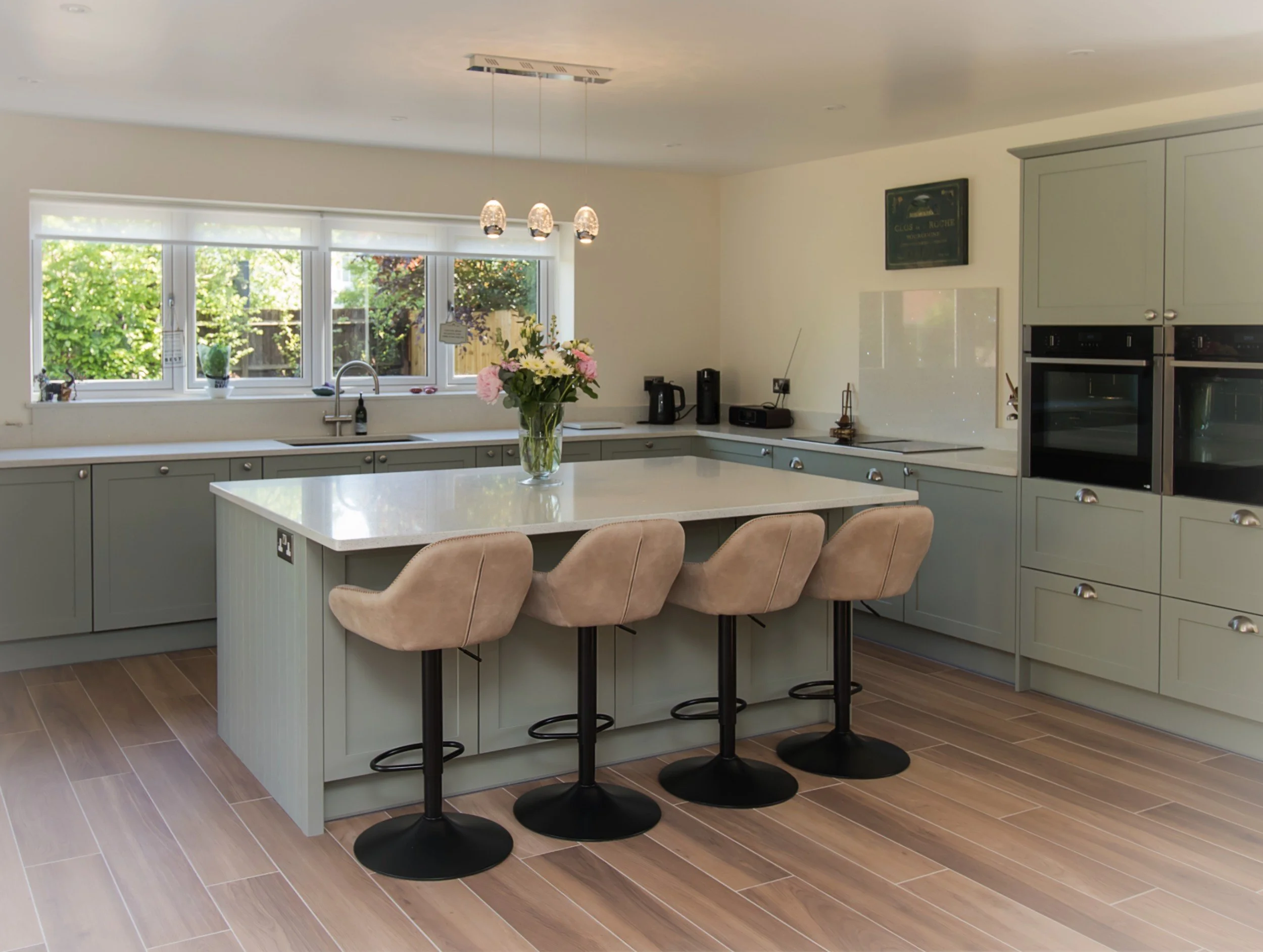 Modern kitchen with light green cabinets, a central island with beige bar stools, a vase with pink and white flowers, and large windows overlooking greenery.