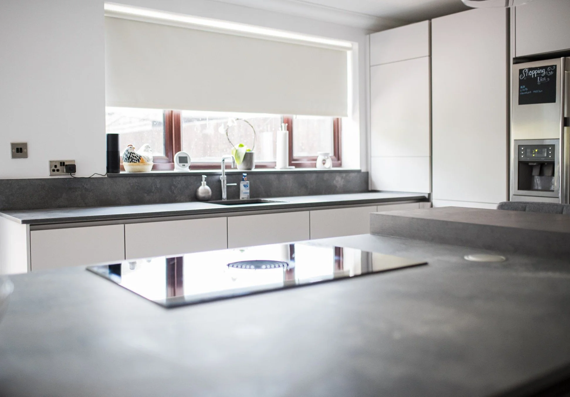 Modern kitchen with white cabinets, gray countertops, a window with a white roller shade, and a stainless steel refrigerator with a chalkboard on the door. On the counter are decorative items, a plant, and small gadgets.