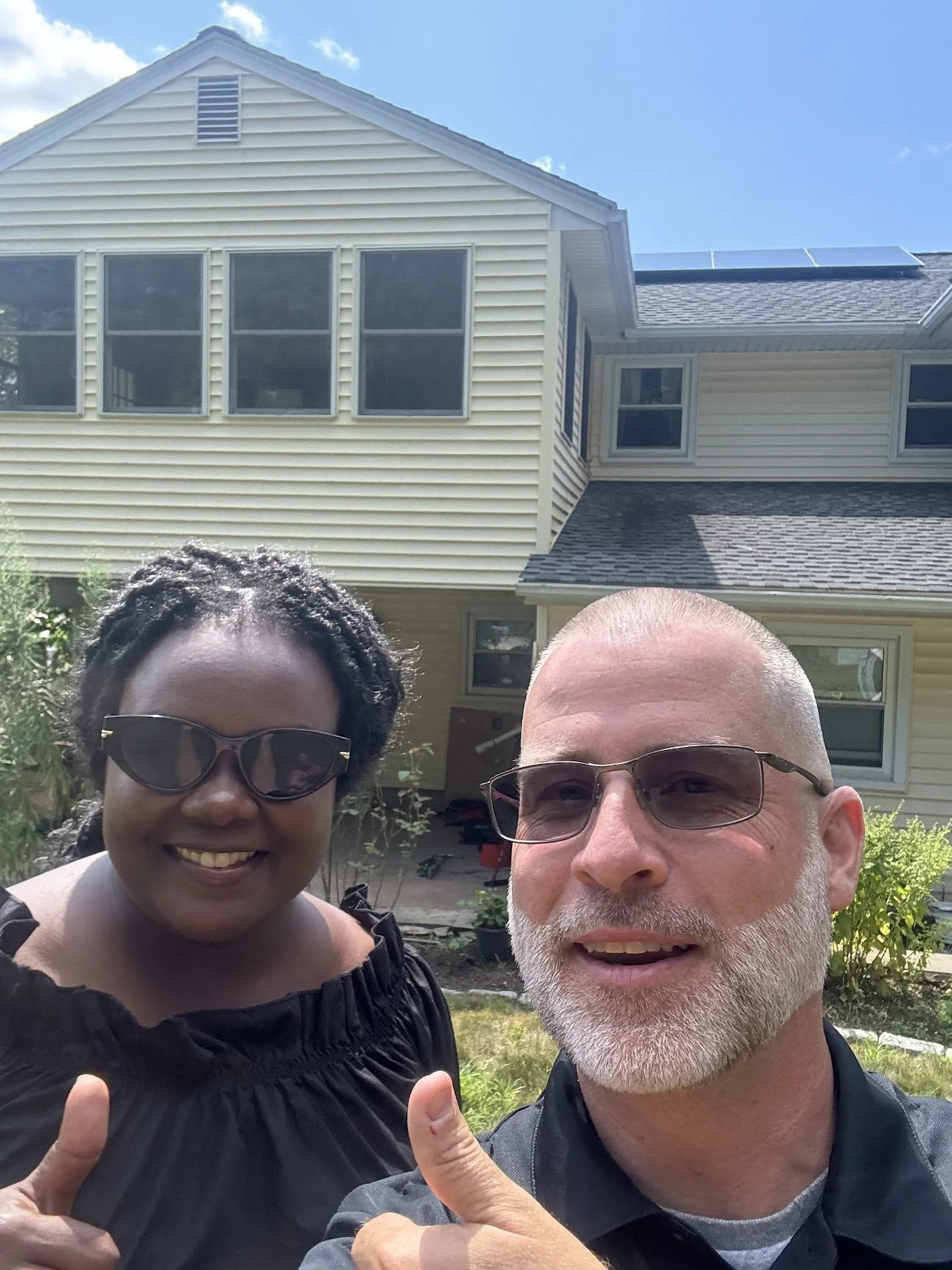 Two people smiling and giving thumbs up in front of a beige house with solar panels on the roof.