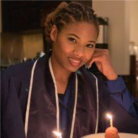 Person smiling next to a cake with lit candles, wearing a blue graduation robe.