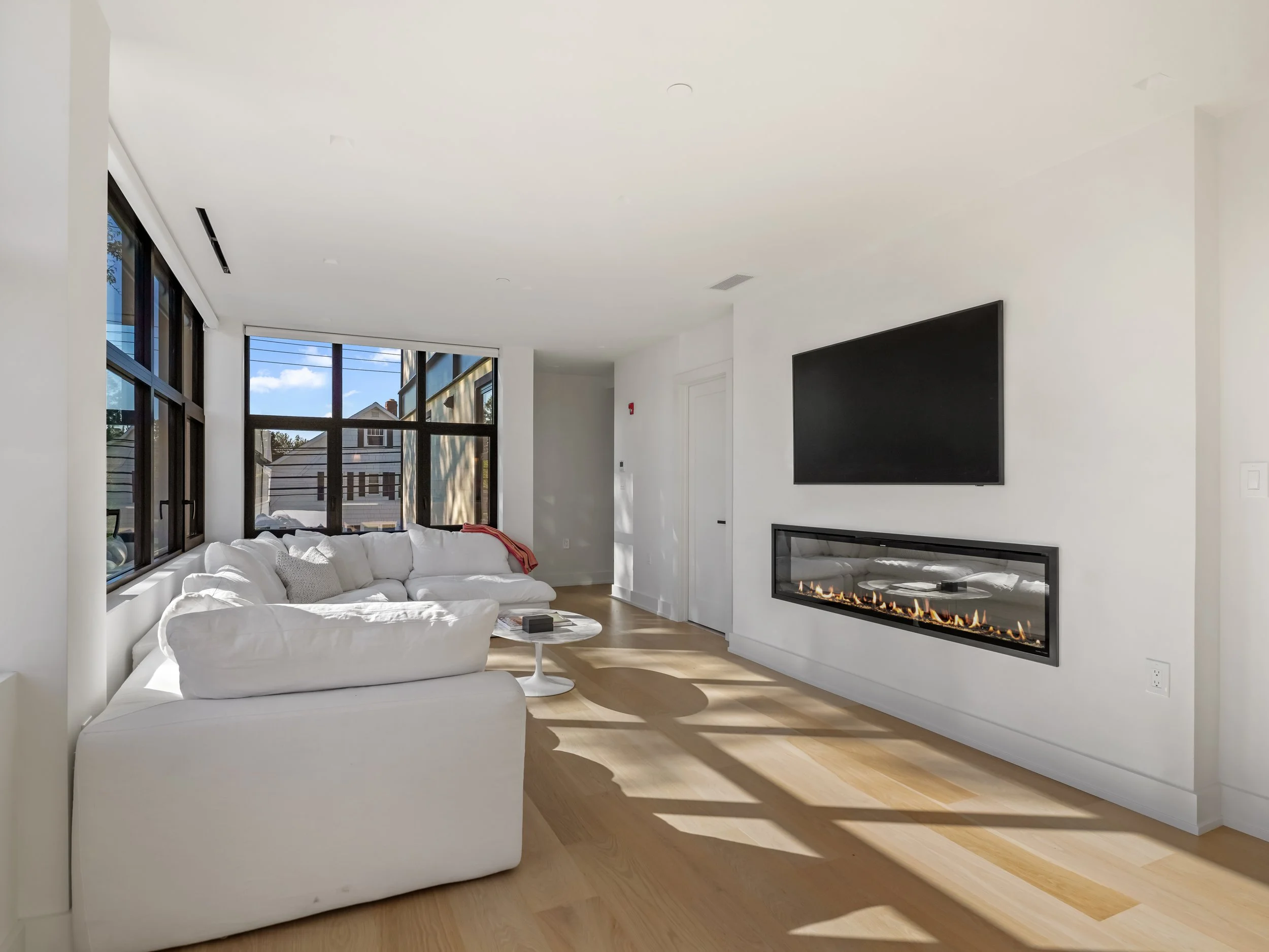 Modern living room with a large white sectional sofa, a small round coffee table, a wall-mounted flat-screen TV, a linear fireplace below the TV, and large windows with black frames showing an outside view.