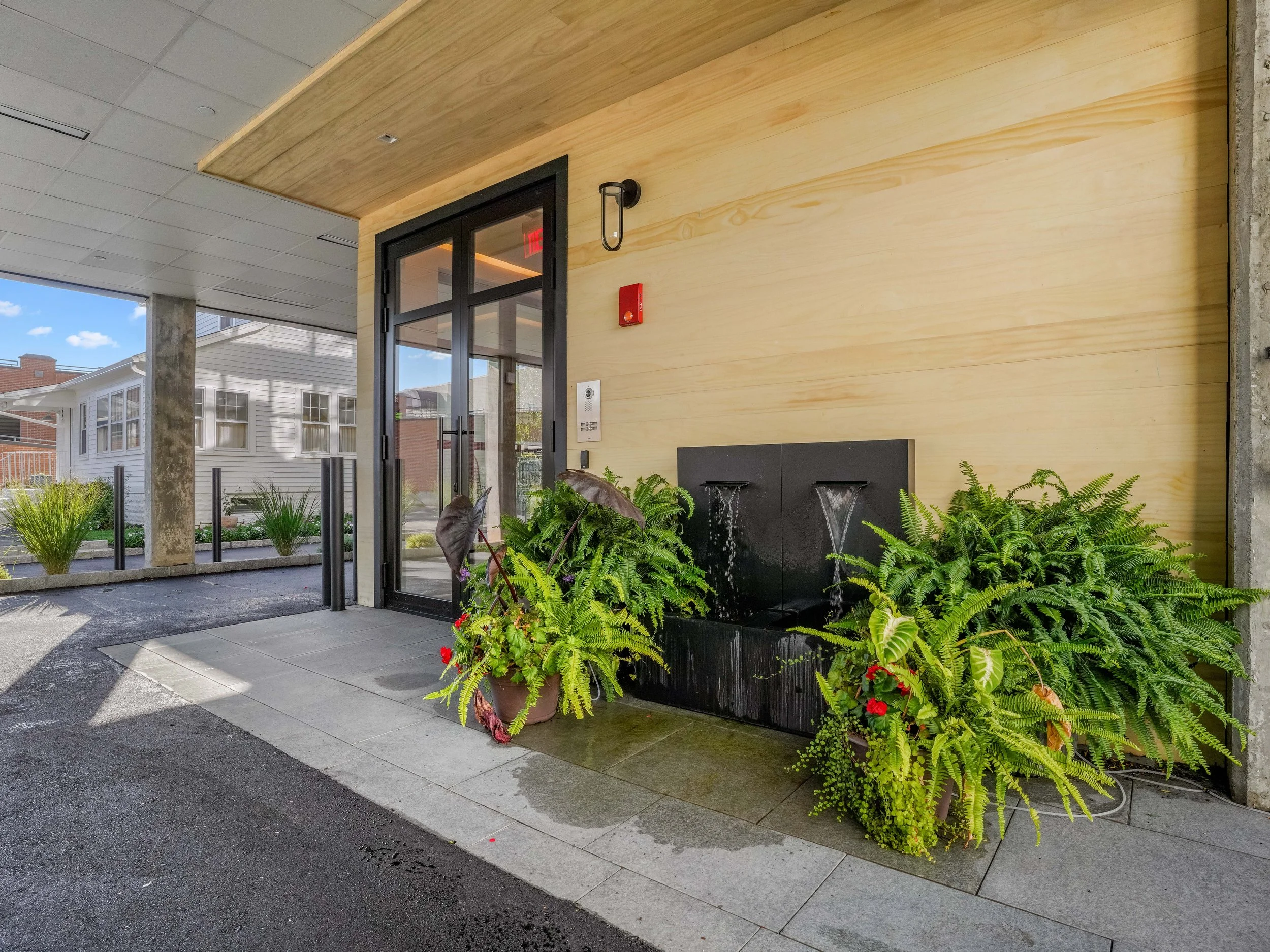 Entrance of a building with glass door, indoor-outdoor water fountain, potted plants, and wooden wall paneling.