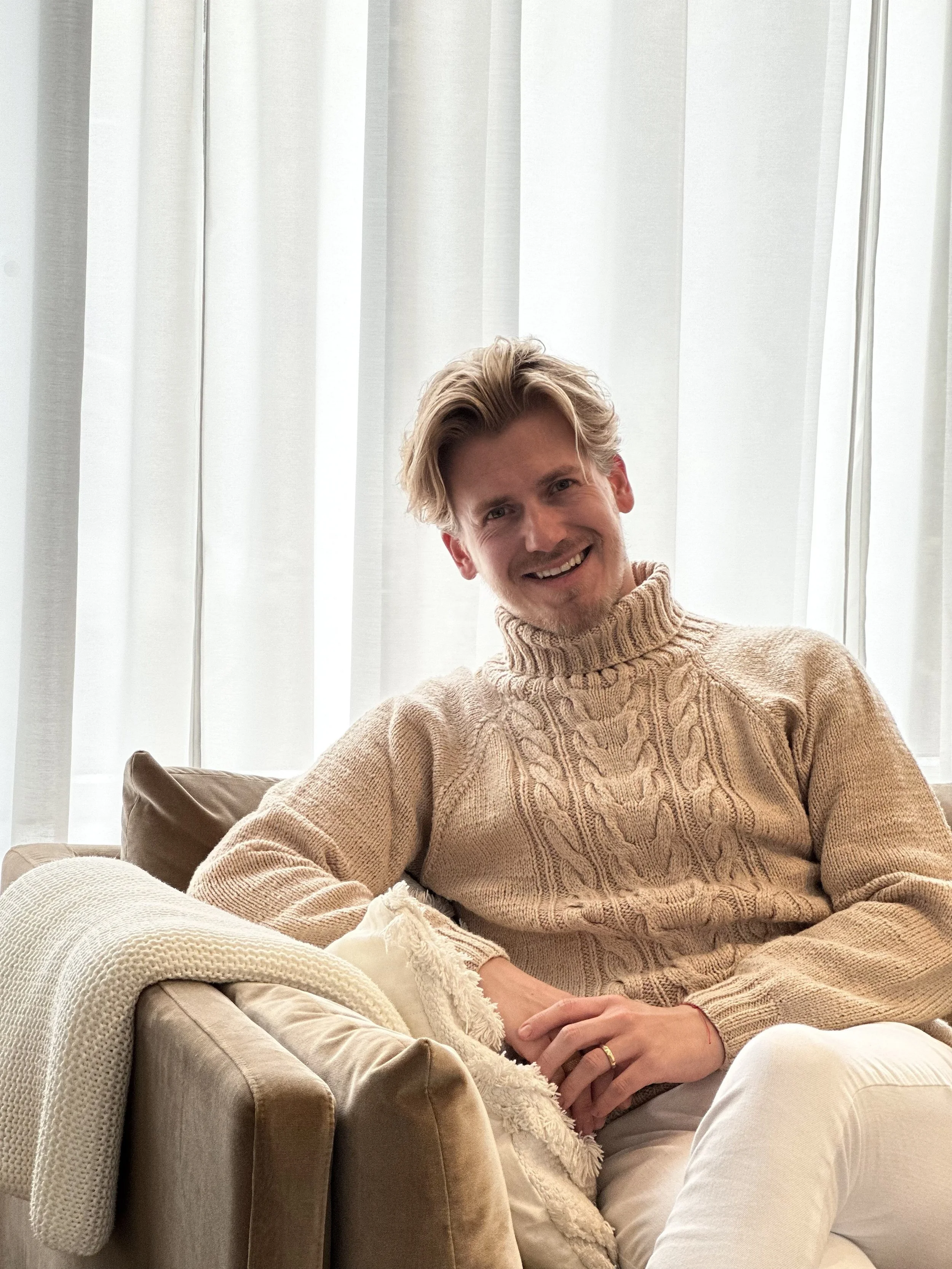 A young man with blonde hair, wearing a beige cable-knit turtleneck sweater, smiling while sitting on a tan sofa in front of white curtains.