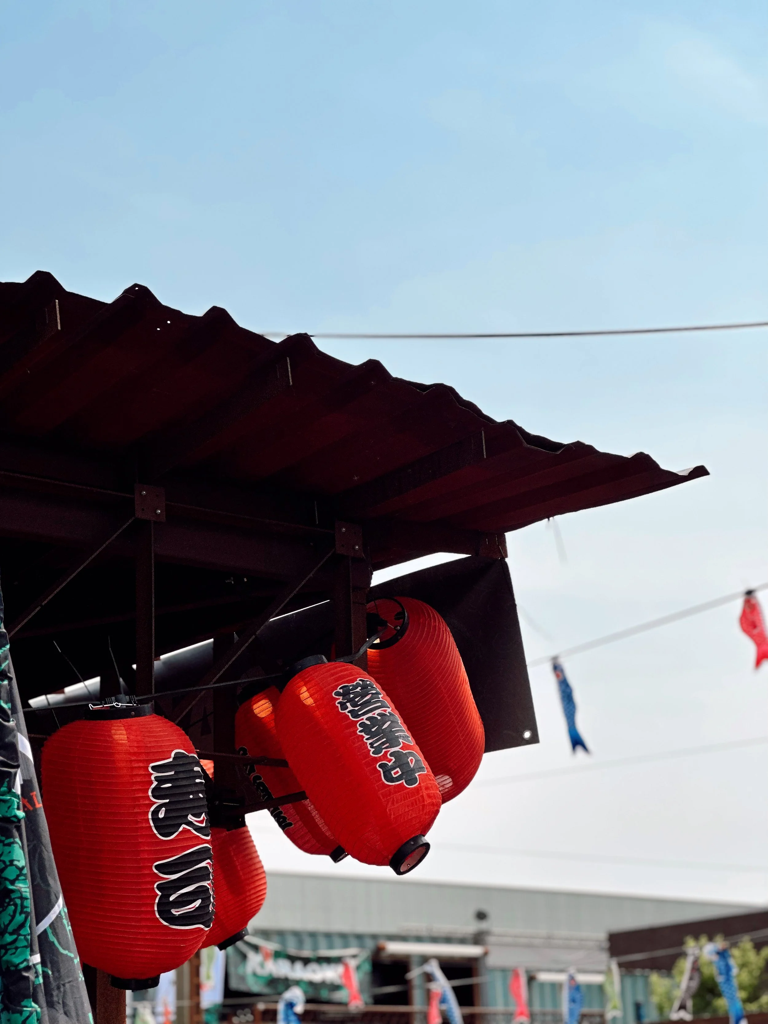 Red paper lanterns hanging under a wooden roof at an outdoor festival with a blue sky and flags in the background.