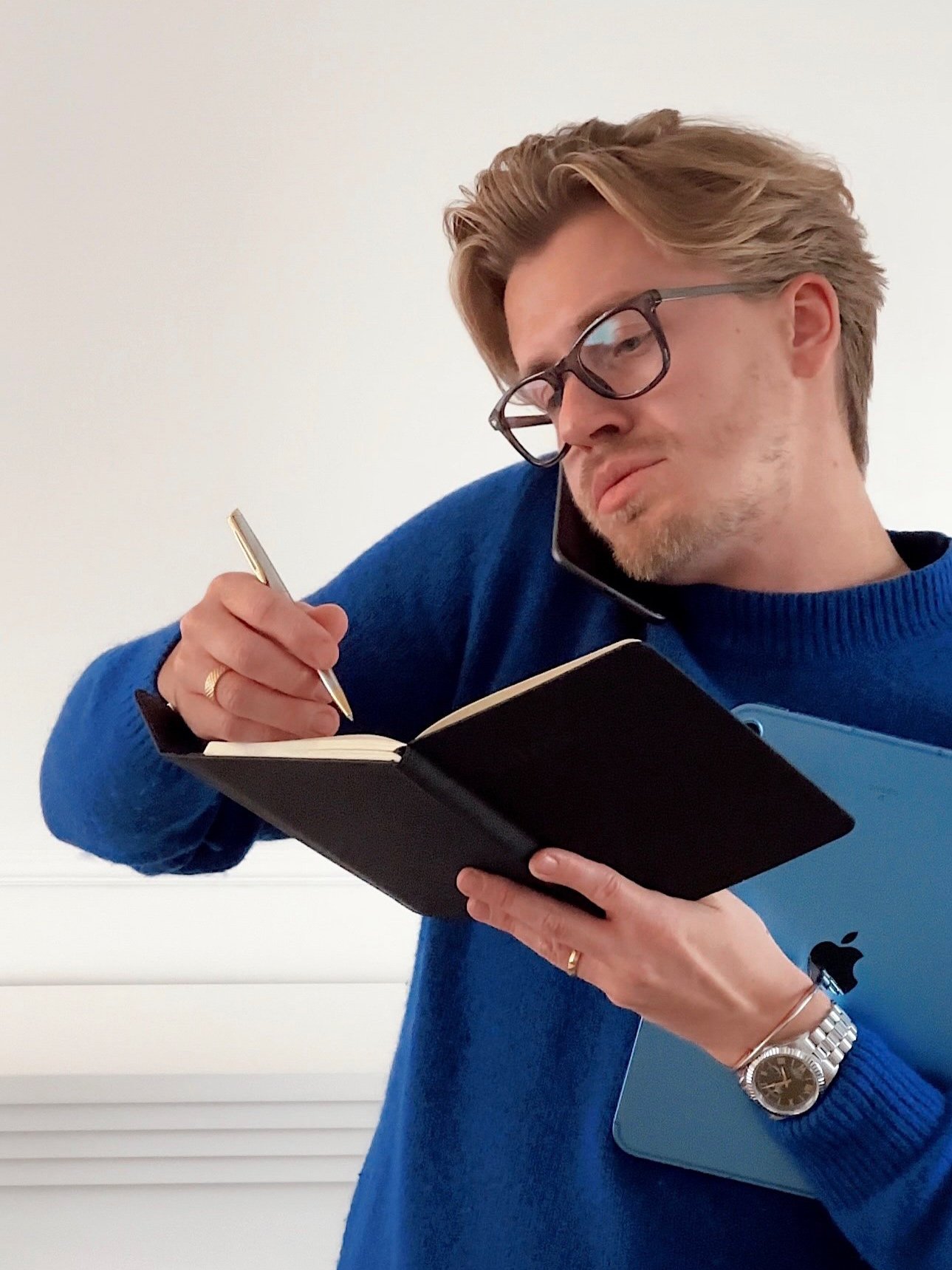 Man with glasses, blue sweater, and watch, writing in a phone book with a pen, holding a blue iPad, talking on a cellphone.