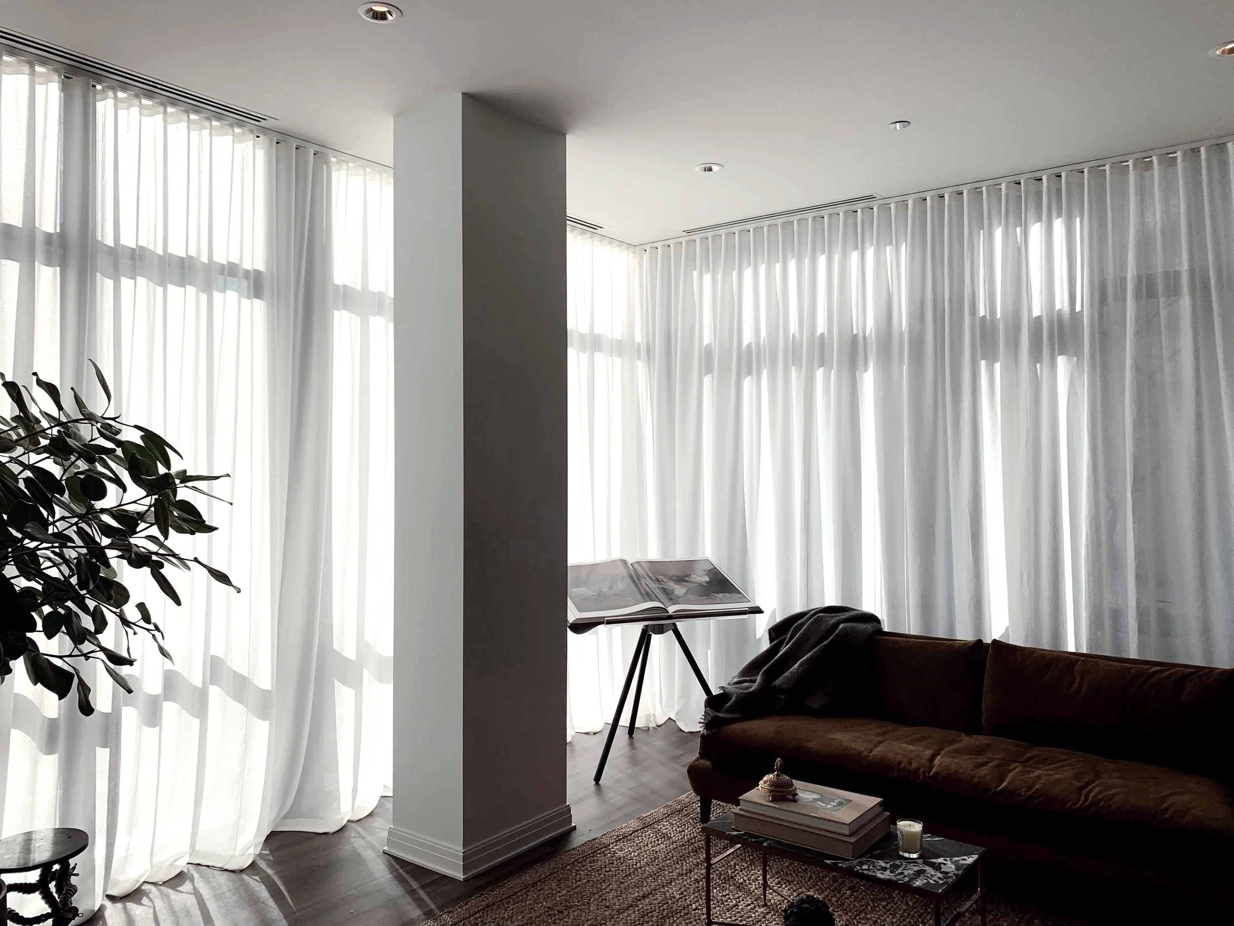 Bright living room with large white curtains, a brown velvet sofa, a coffee table with books, a small decorative object, a candle, an art book stand, and a potted plant.