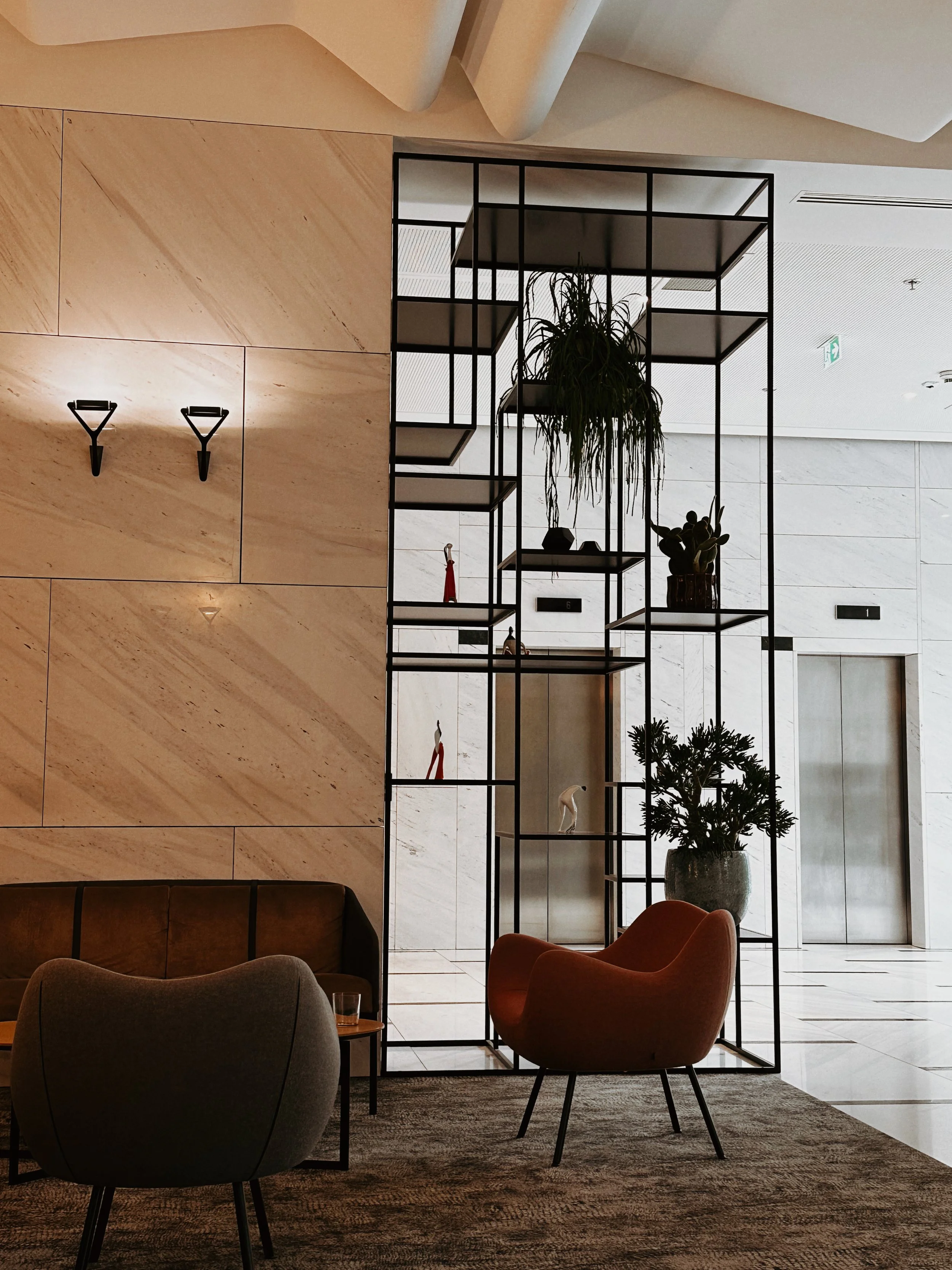 Interior of a modern lobby or lounge area with a black metal and glass display room, decorative plants inside, beige marble walls, and contemporary furniture, including a gray and an orange armchair.