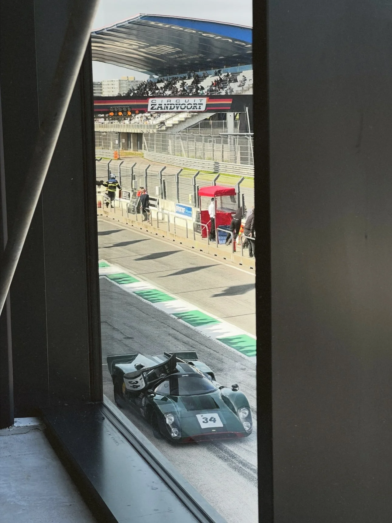 A vintage race car with the number 34 on the hood and the number 33 on the side is parked in a pit lane at a racetrack. The photo is taken from inside a building, looking through a window at the car and the pit area with people and equipment around.