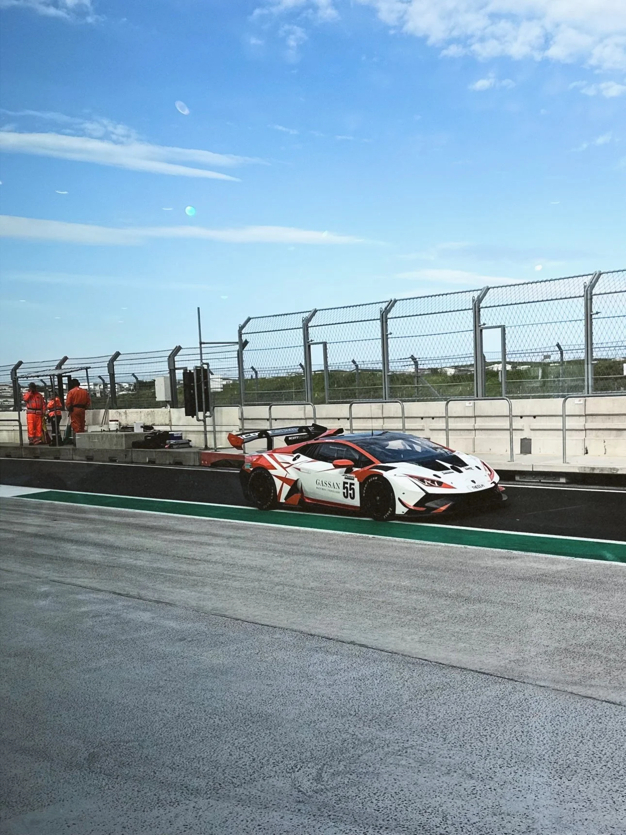 A white race car with red and black accents parked in a pit stop area at a race track, with pit crew members in orange uniforms standing nearby under a blue sky.