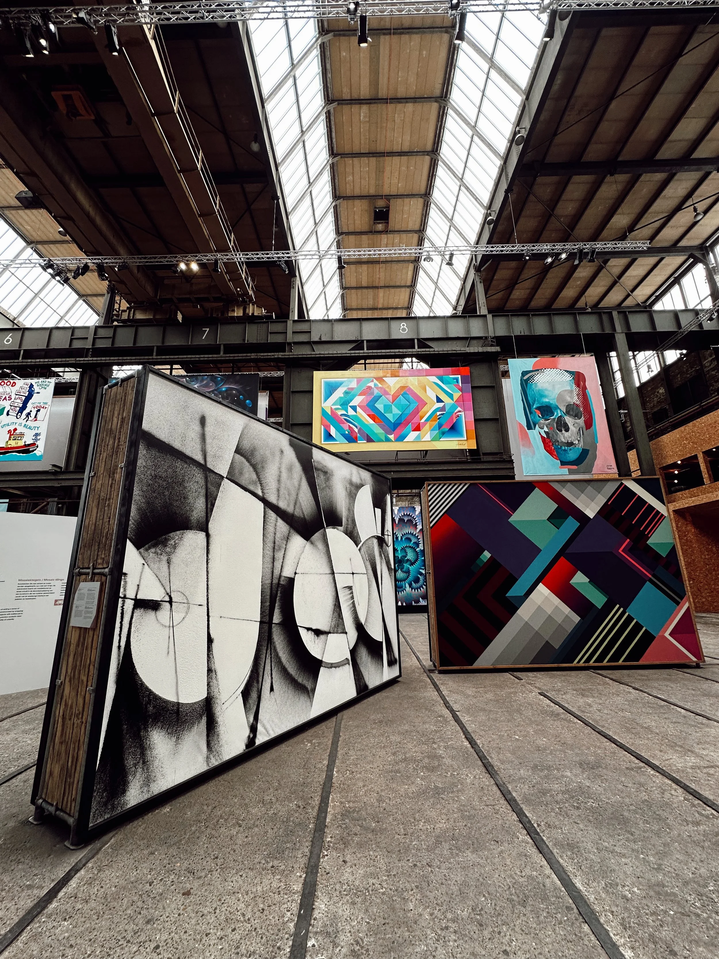 Art exhibition inside a large industrial-style building with a high, glass ceiling. There are several colorful and monochrome paintings and paintings on display, including geometric patterns, a skull with colorful accents, and abstract designs.