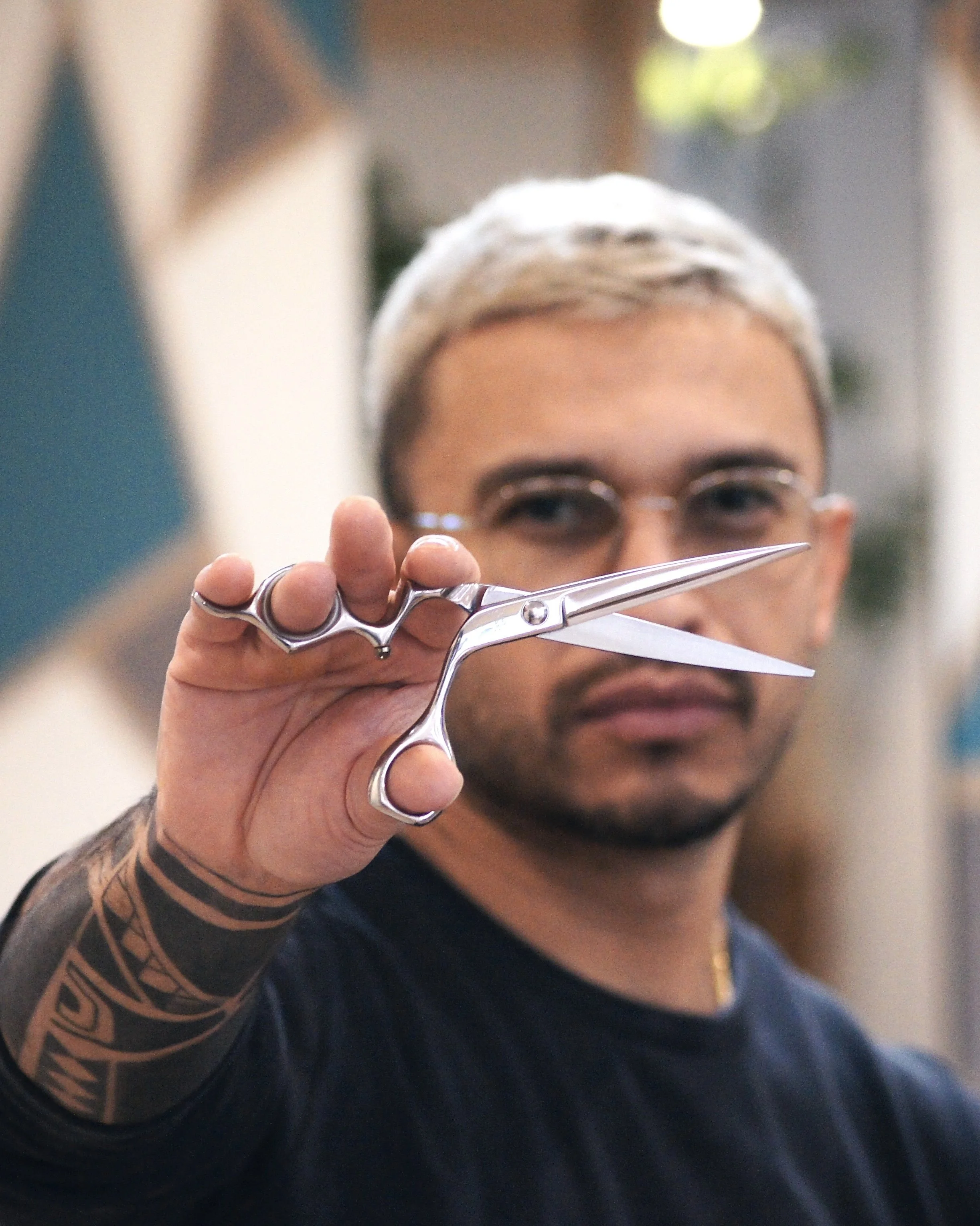 A person with short, bleached hair and glasses holding a pair of silver scissors towards the camera, with a blurred background.