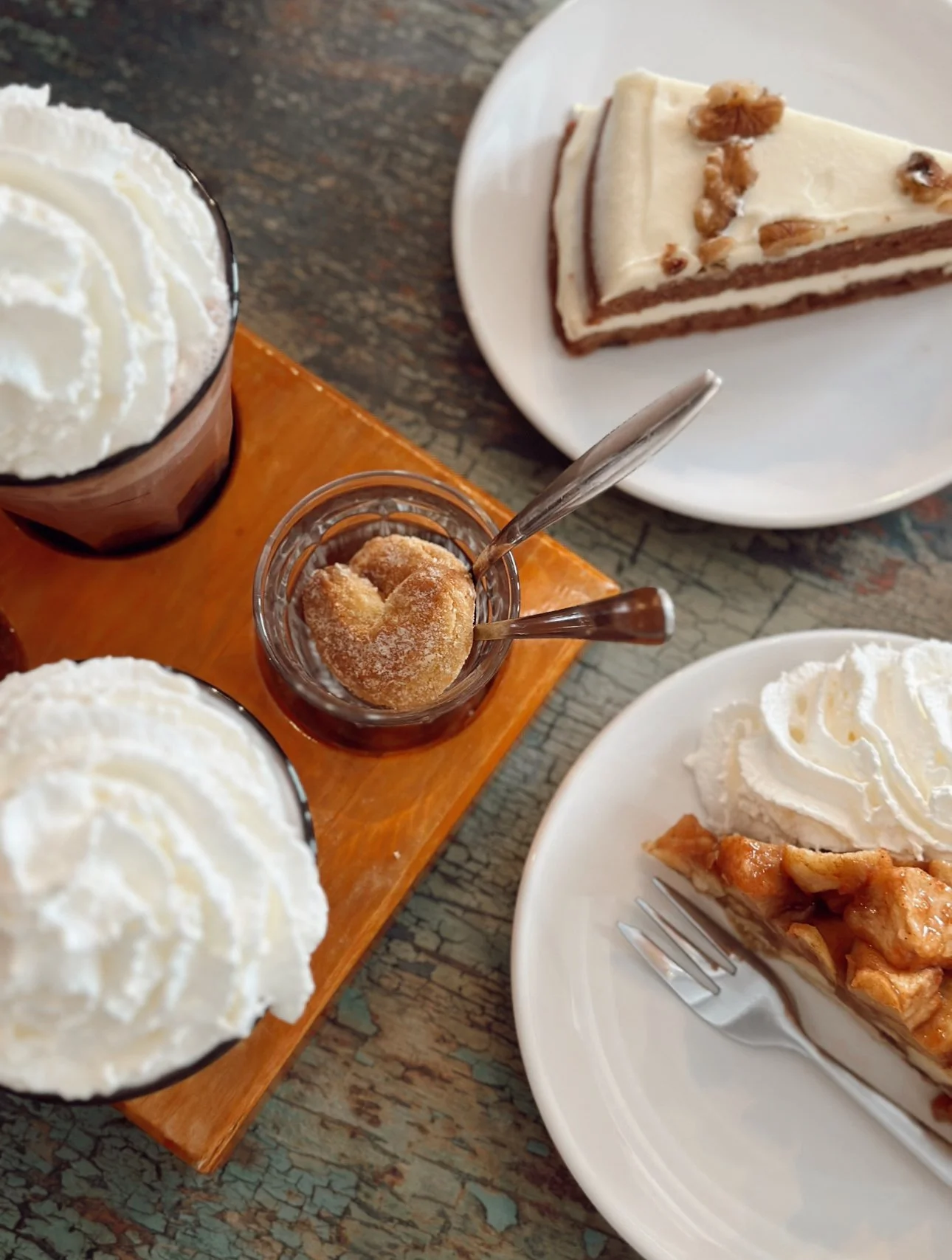 Two glasses of whipped cream-topped beverages, a small jar with a heart-shaped pastry in syrup, a slice of layered cake with white icing and walnuts, and a slice of apple pie with whipped cream on a white plate, all on a rustic wooden table.