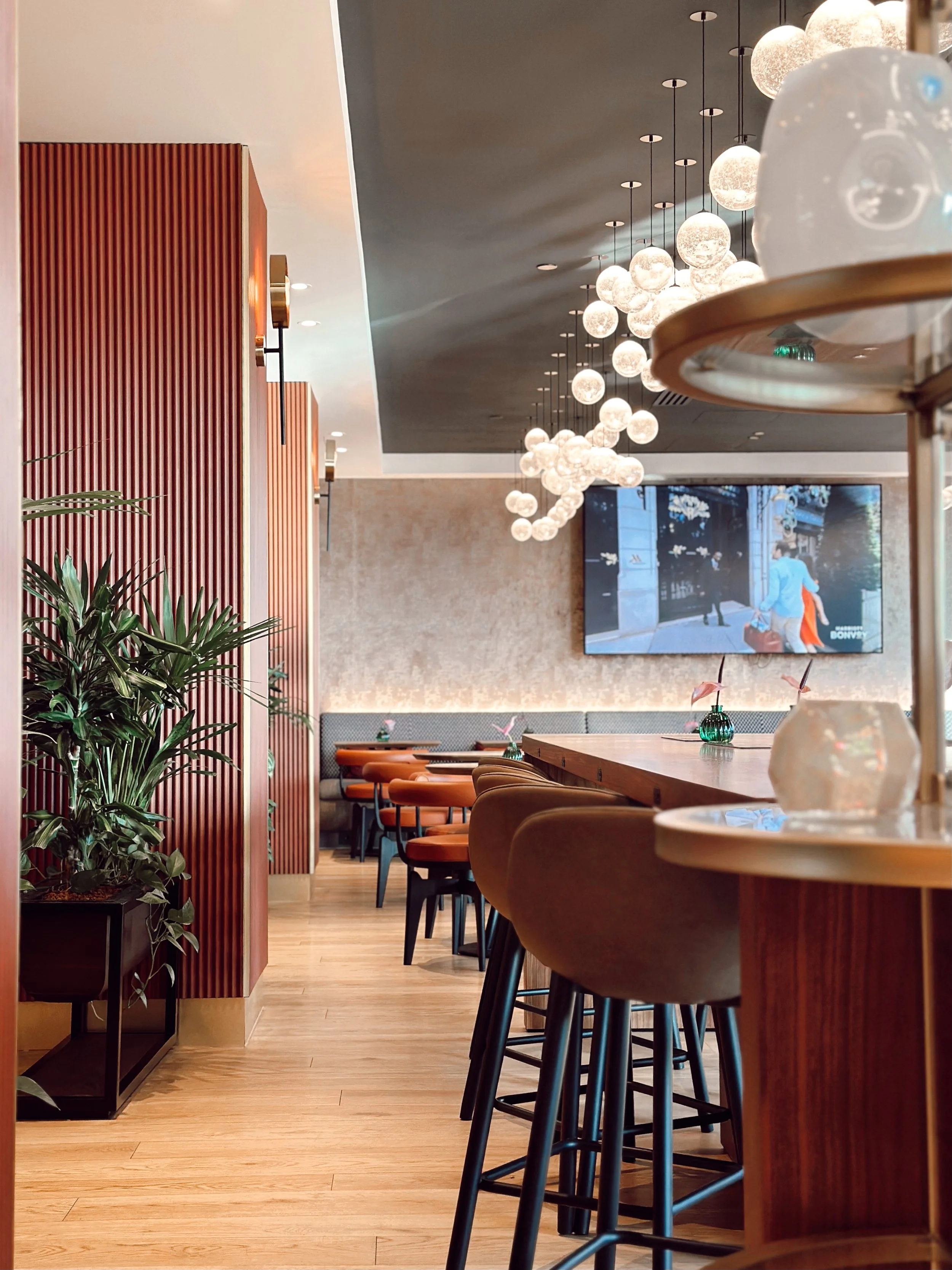 Modern restaurant interior with wooden accents, hanging spherical light fixtures, bar stools, a long wooden bar counter, a potted plant, and a wall-mounted TV screen