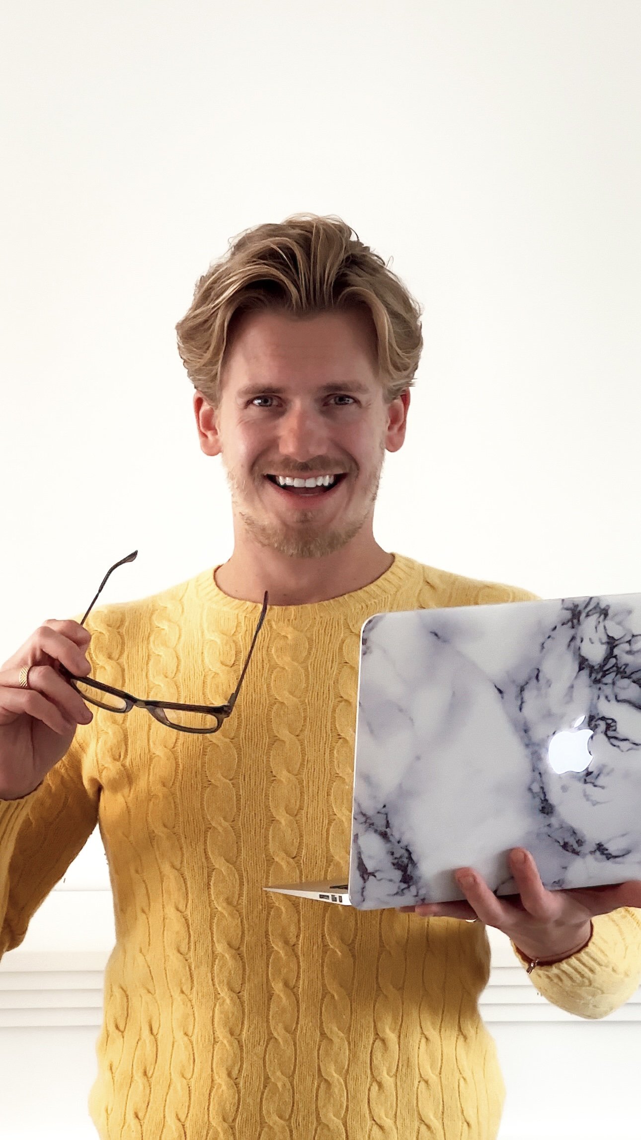 A young man with blond hair and a beard smiles, holding a marble-patterned MacBook in one hand and a pair of glasses in the other, standing against a plain white background.