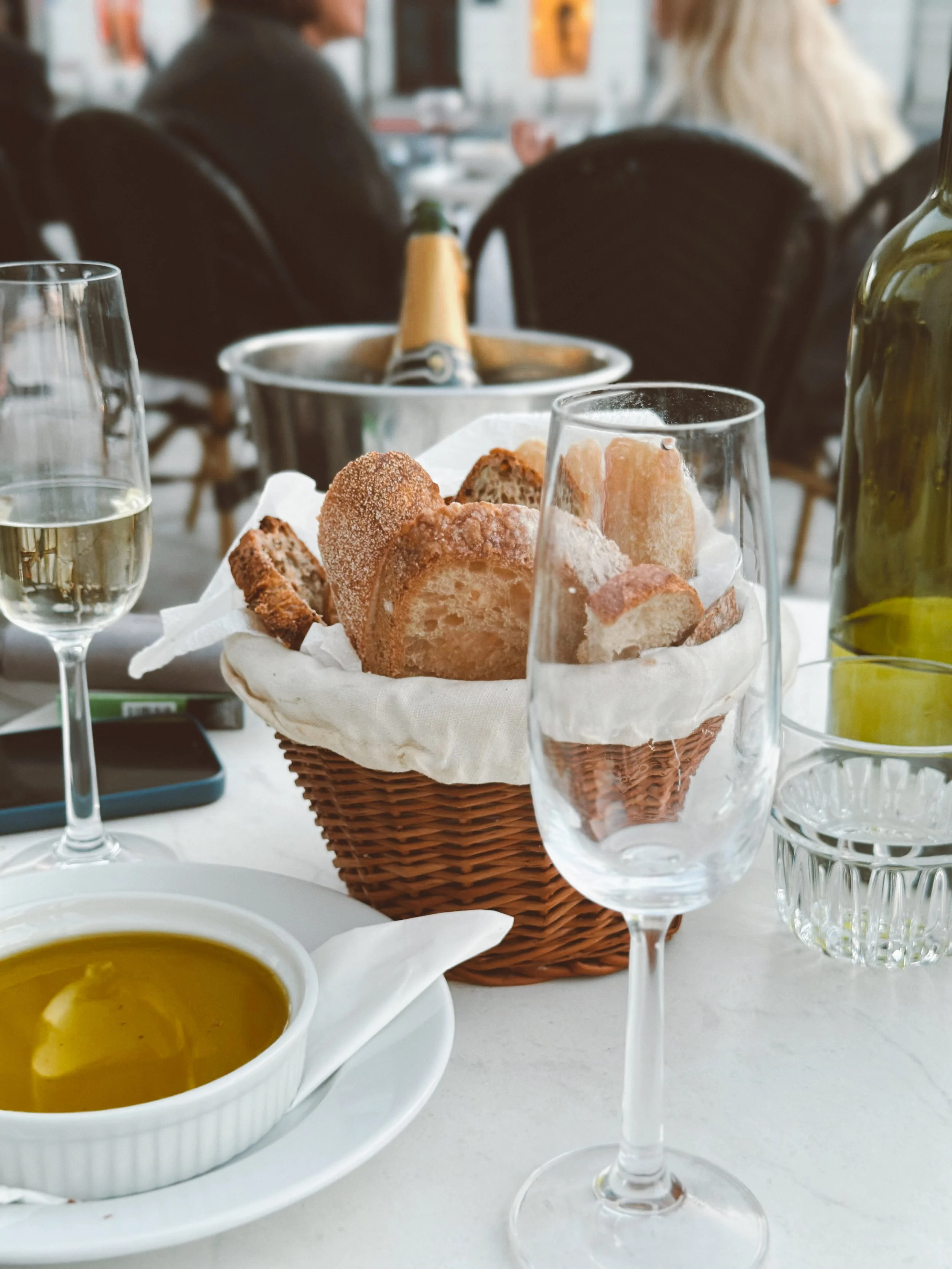 Basket of sliced bread, two glasses of white wine, a small bowl of olive oil, and a glass of water on a dining table at a restaurant.