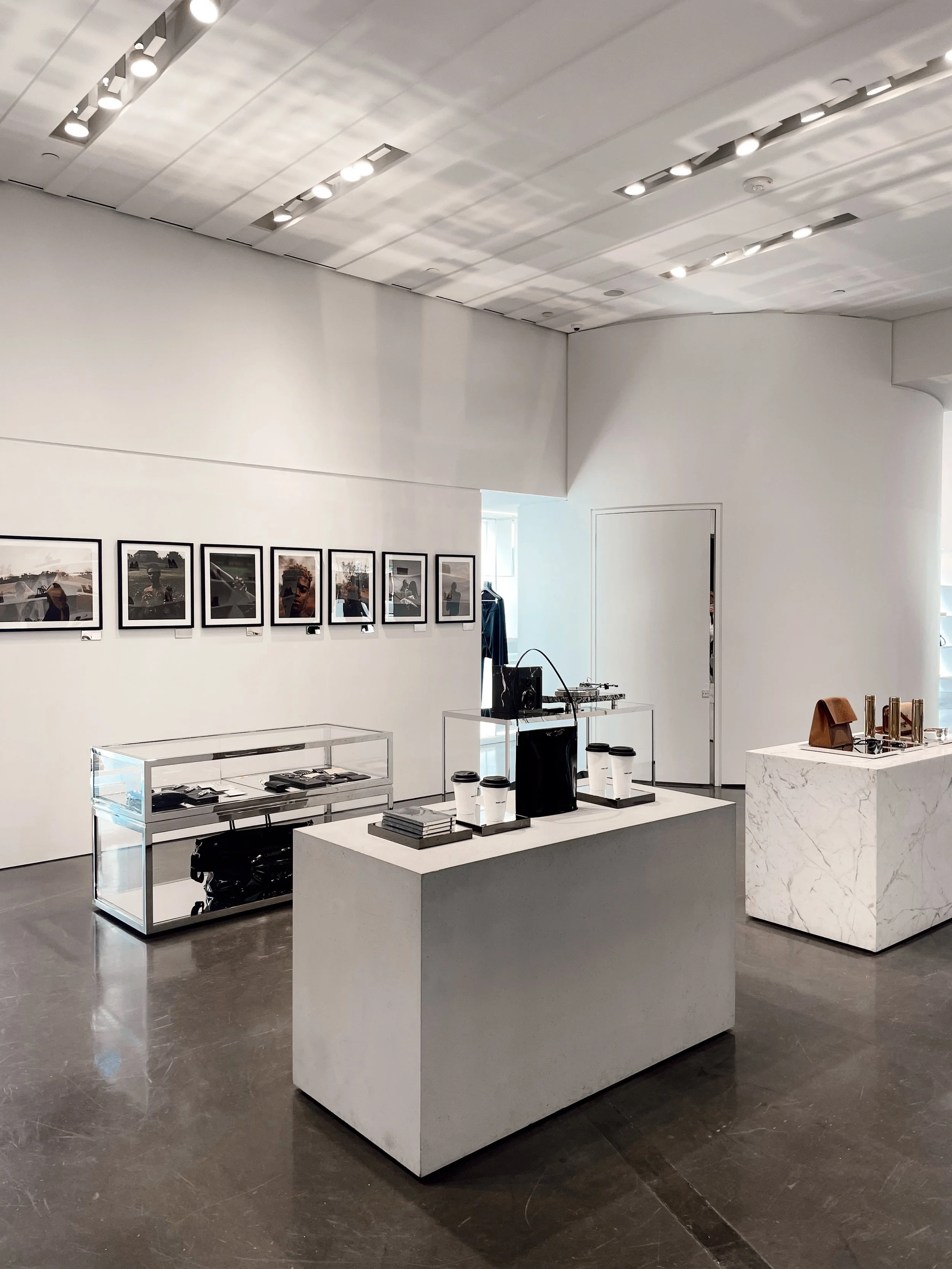 Interior of a modern retail store with white walls and ceiling, featuring framed photographs on the wall. Display tables hold various accessories including handbags, books, cups, and jewelry.