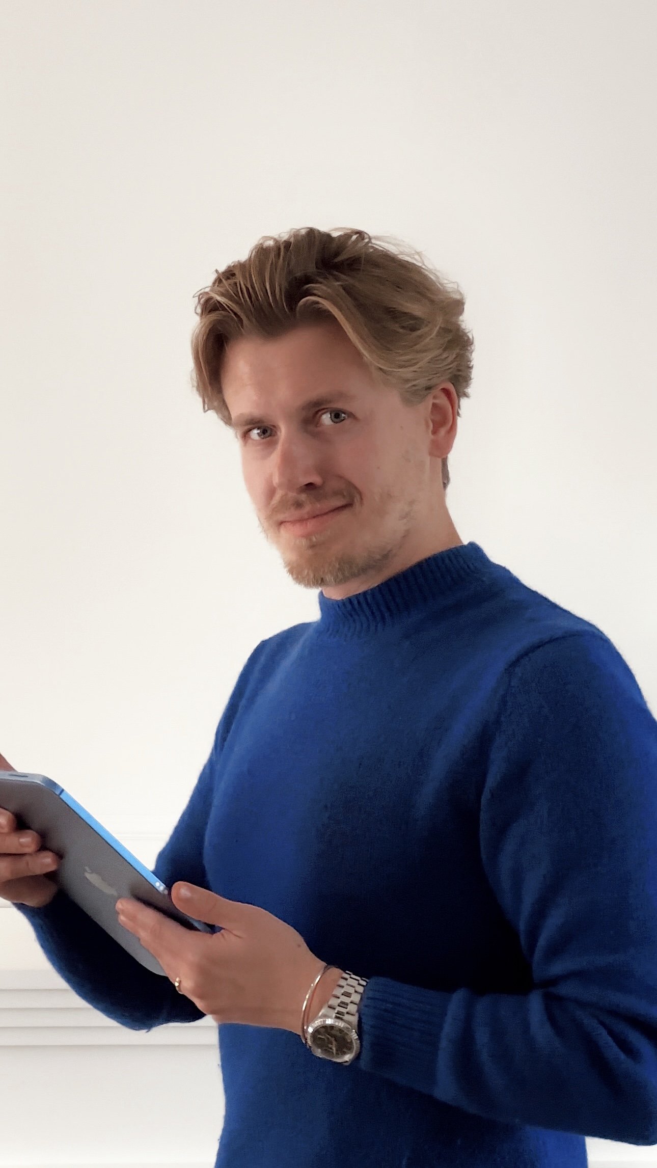 Man with blond hair wearing a blue sweater, holding a smartphone, looking at the camera, standing against a plain white wall.