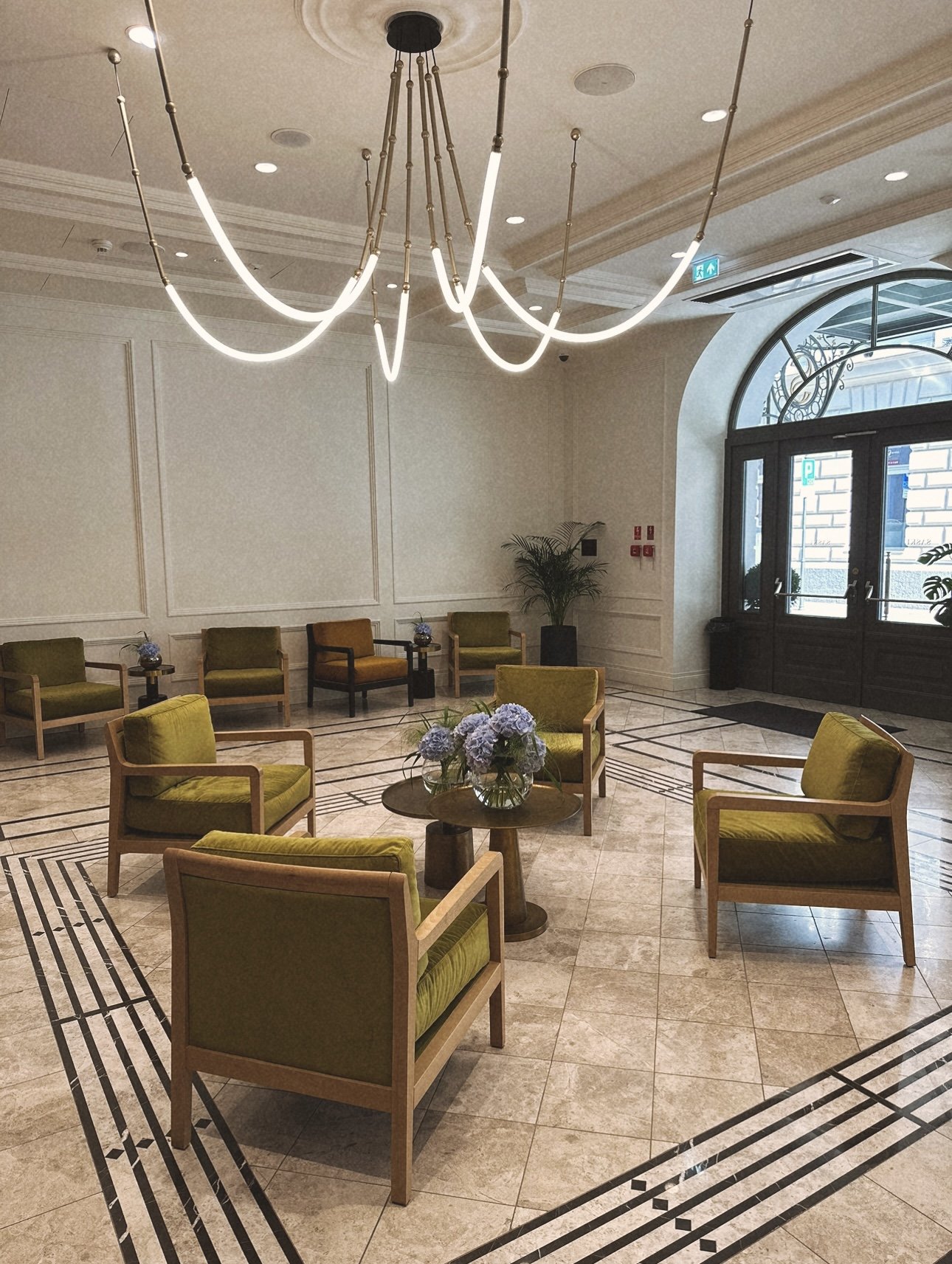 A spacious lobby with a modern chandelier hanging from the ceiling, arranged with green and mustard-colored armchairs around wooden tables, some with flower arrangements, and a large arched window allowing natural light, along with potted plants and 