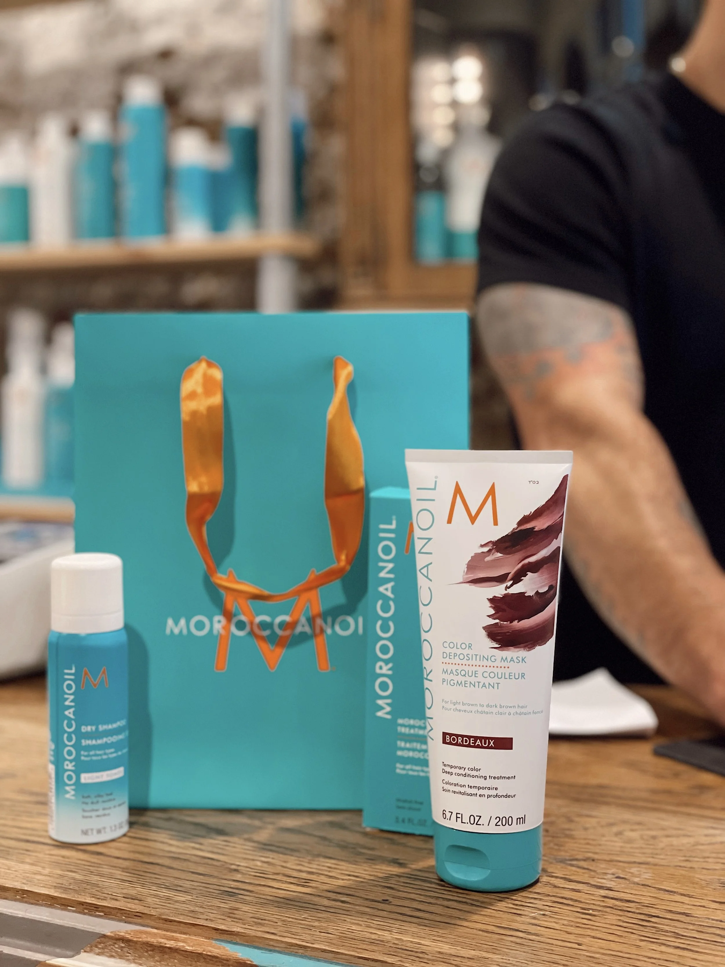 Moroccanoil hair care products, including a blue bottle, a tube with a maroon design, and a blue paper bag with orange handles, placed on a wooden surface.