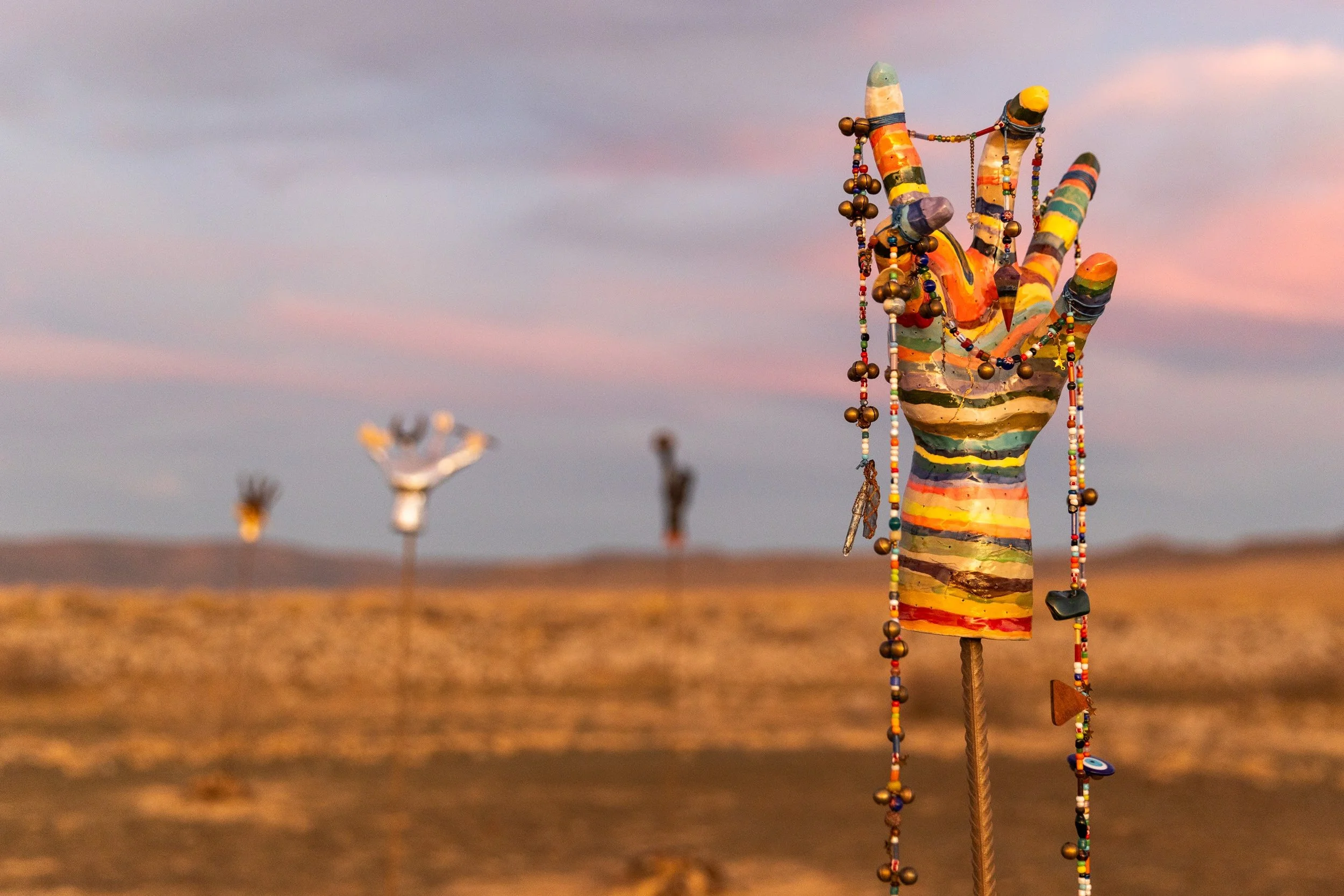 Colorful, abstract sculpture of a hand with beads and jewelry hanging from it, in a desert landscape during sunset with metal sculptures in the background.