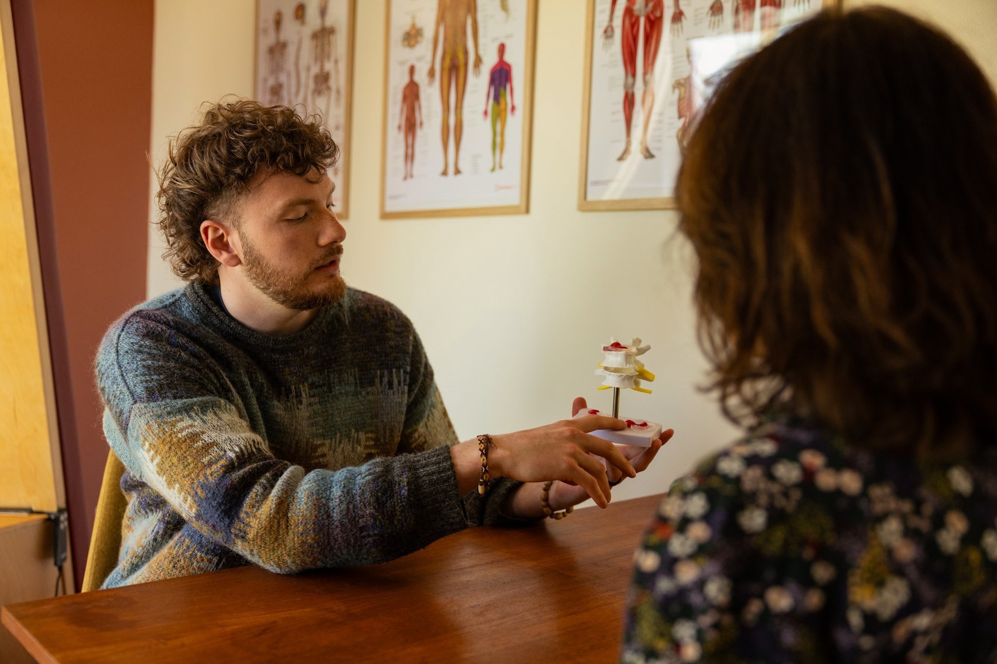 Twee mensen zitten aan een tafel bij BSR Mandala. Eén persoon houdt een model van een menselijke wervelkolom vast en legt het mogelijk aan de ander uit. Er hangen anatomische posters aan de muur. Beide personen zijn gekleed in casual kleding.