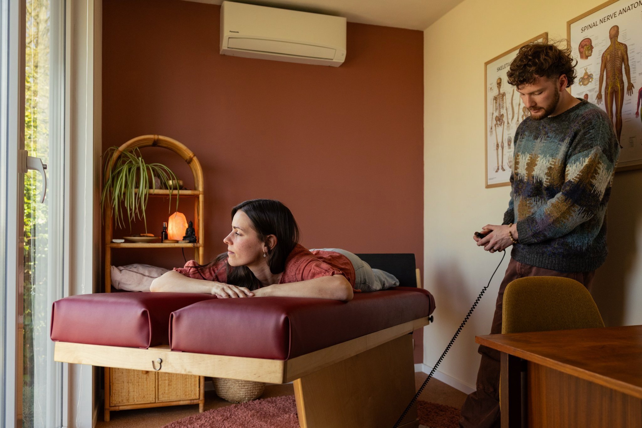 Foto van Lennert, Body Stress Release practitioner bij BSR Mandala, die een client op de behandelbank omhoog zet met een afstandsbediening. De kamer heeft een warme inrichting met planten en een raam dat daglicht binnenlaat.