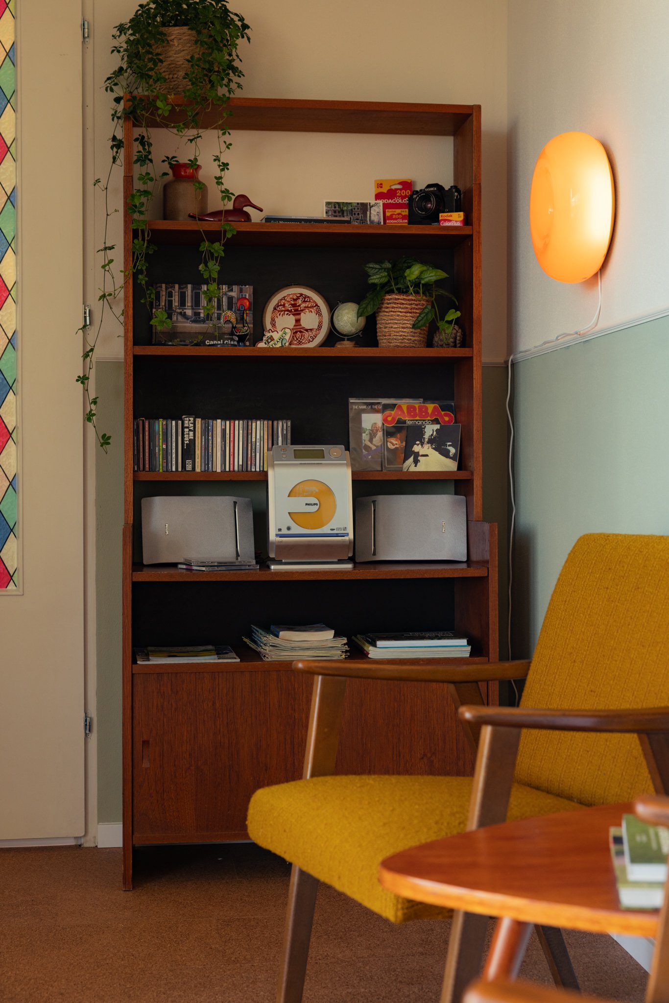 Interieur met retro-stijl plank met boeken, cd's, planten; naast een gele stoel en ronde lamp aan de muur.