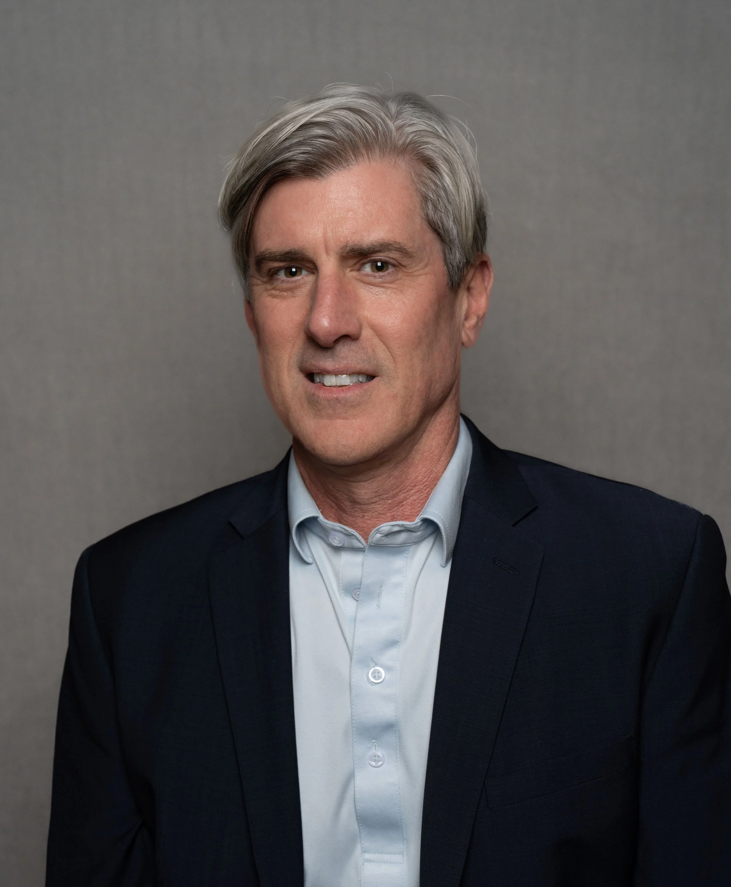 A middle-aged man with gray hair, wearing a dark suit jacket over a light blue shirt, smiling at the camera against a plain gray background.