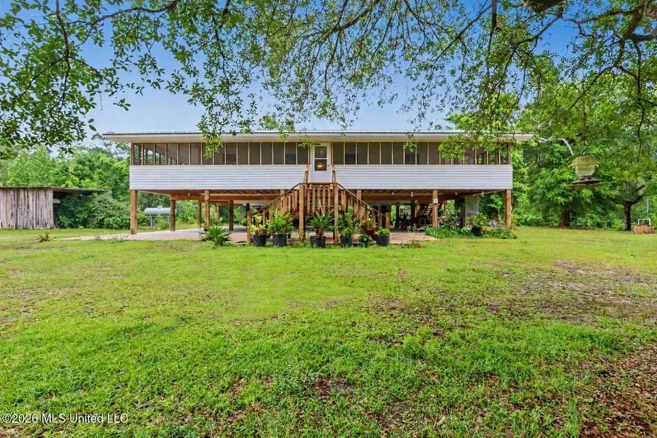 Home for sale in Lucedale. Home for sale in George County. Home on the river in Lucedale with a raised porch and stairs, surrounded by a grassy yard and trees, with a small shed on the left and bird feeders hanging from trees.
