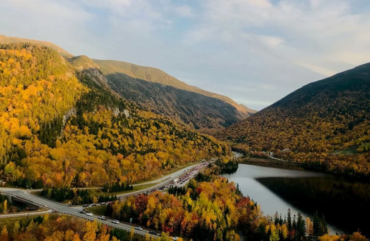 A scenic view of a lake surrounded by mountains with trees in fall colors and a winding road near the water.
