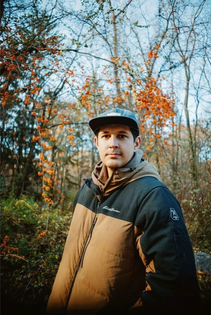 A young man in outdoor clothing, including a cap and a brown and black jacket, stands in a wooded area with trees and orange autumn leaves in the background.