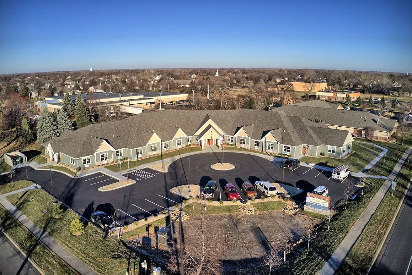 Aerial view of a residential senior care facility with a parking lot and surrounding trees, in a suburban area.